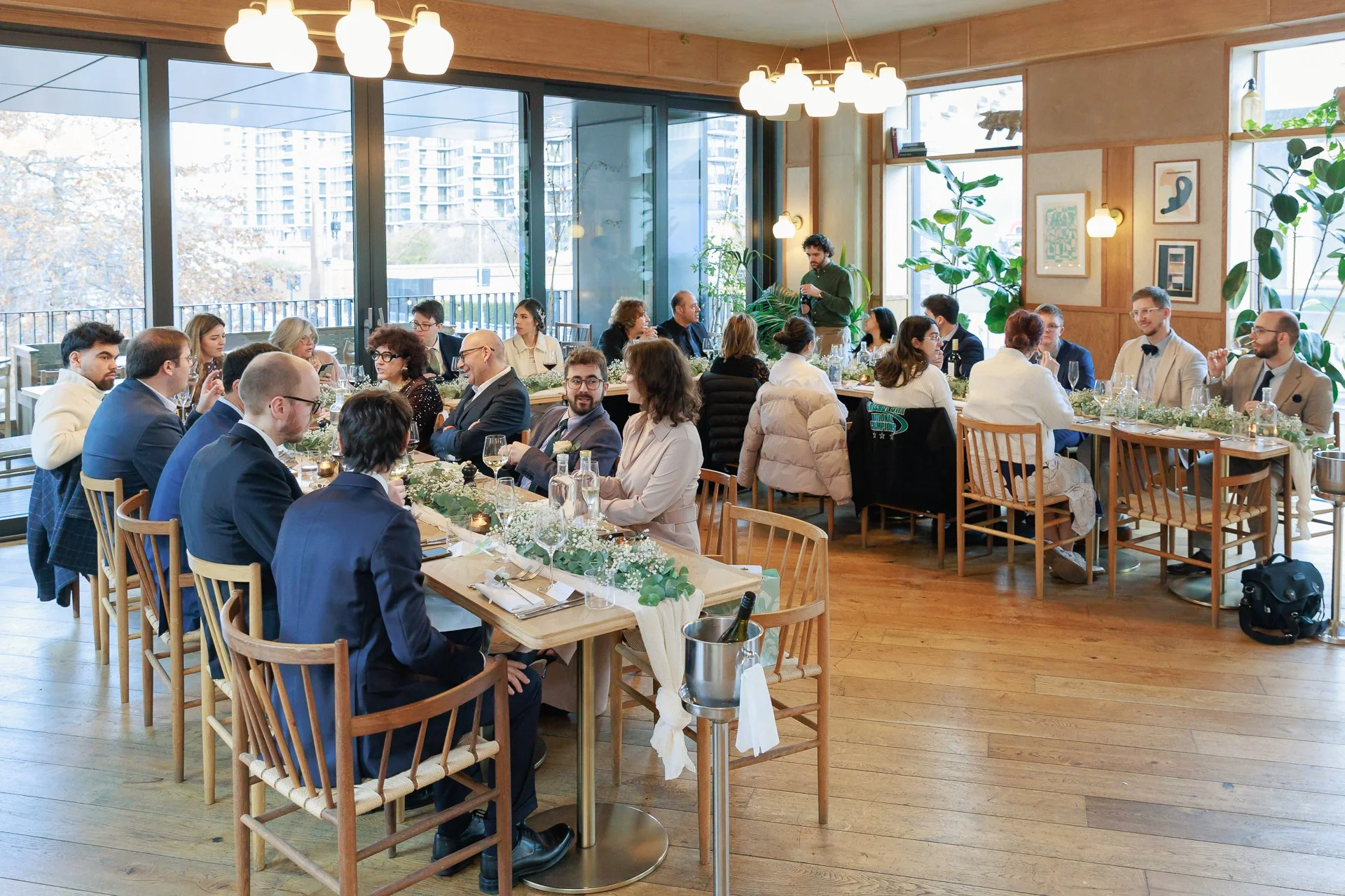 A large group of people dressed in formal attire sitting around a rectangular dining table with elegant decorations in a well-lit restaurant or event space with large windows and indoor plants.