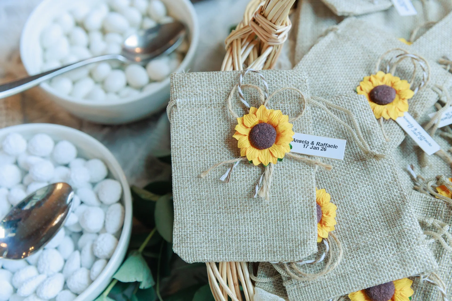 Small booklet on a basket with a sunflower-shaped decoration, surrounded by more wrapped packages with sunflower decorations, and bowls of white candies with spoons.