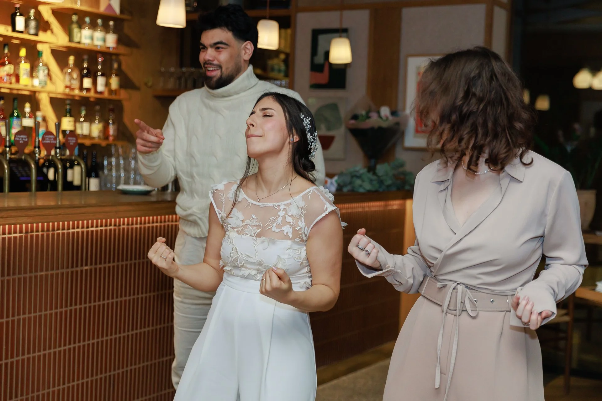 Three people at a bar, two women and one man, celebrating with smiles and clenched fists. The woman in the center is wearing a white dress with lace detailing, and the woman on the right is dressed in a beige outfit. The man in the background is wear