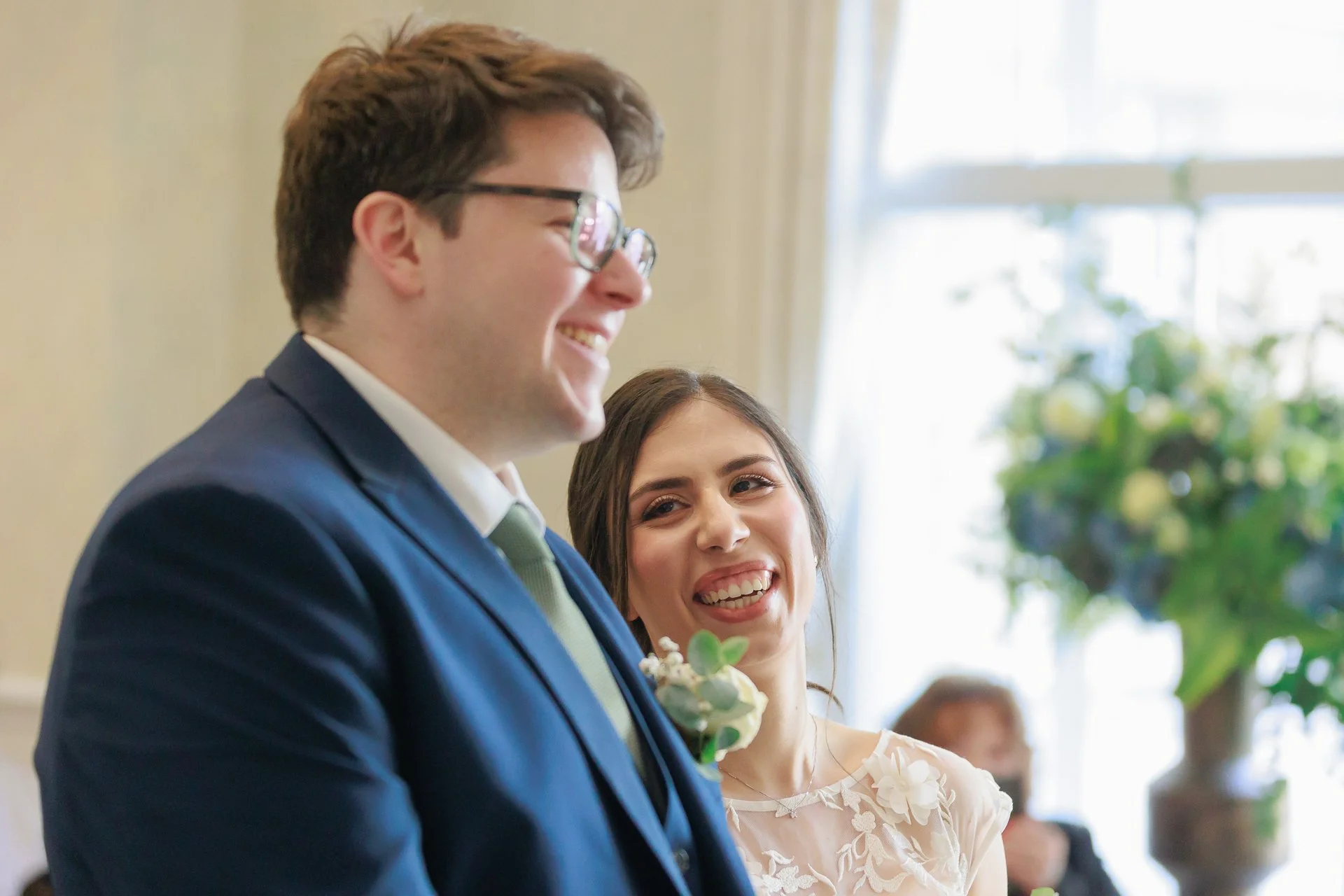 A groom in a dark blue suit and glasses smiling, standing next to a bride in a white floral dress at a wedding ceremony.