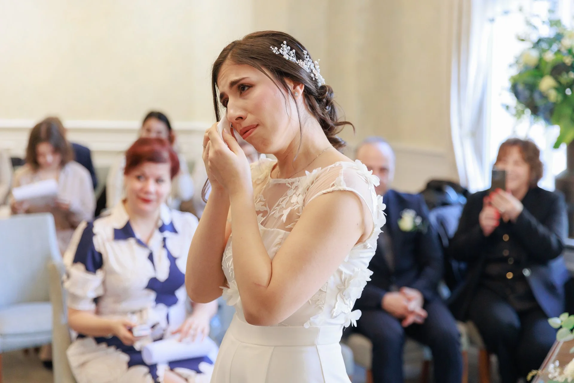 A bride crying, holding a tissue to her face during her wedding ceremony, with guests in the background reacting emotionally.