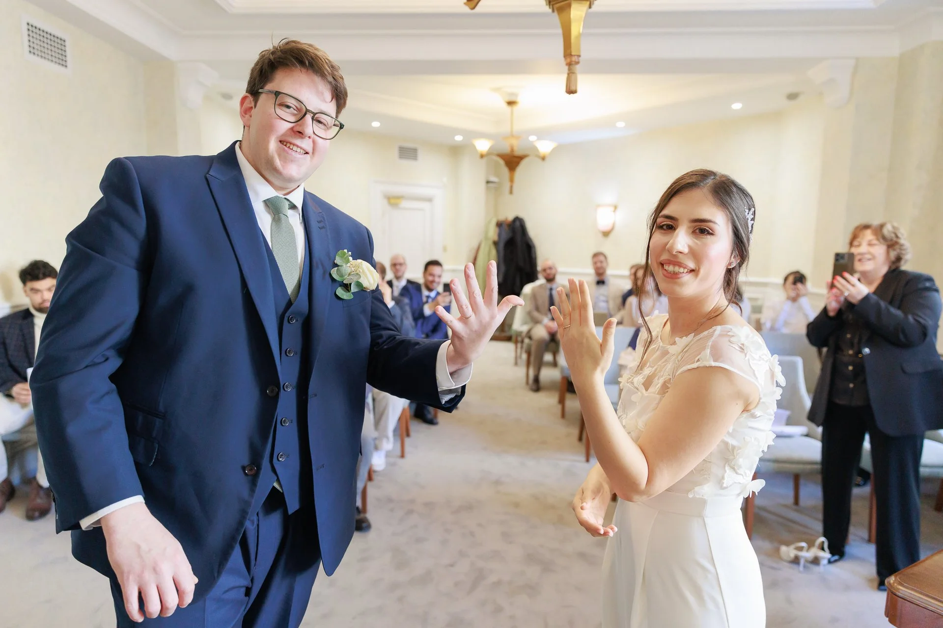 A groom and bride showing their wedding rings during a wedding ceremony, with guests in the background.