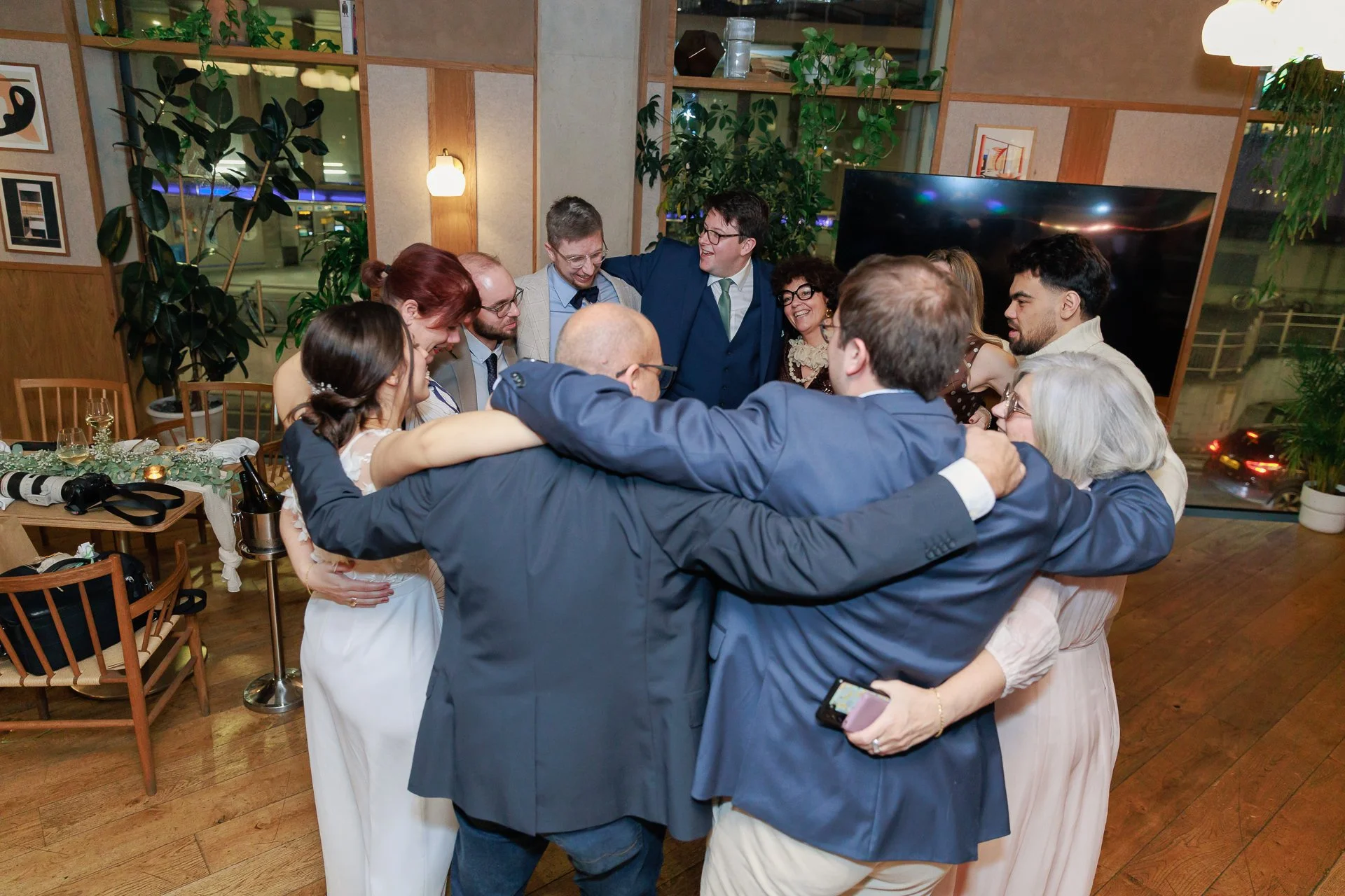 Group of people in formal attire embracing in a circle at a celebration or wedding reception in a warmly decorated room with green plants and wooden furniture.