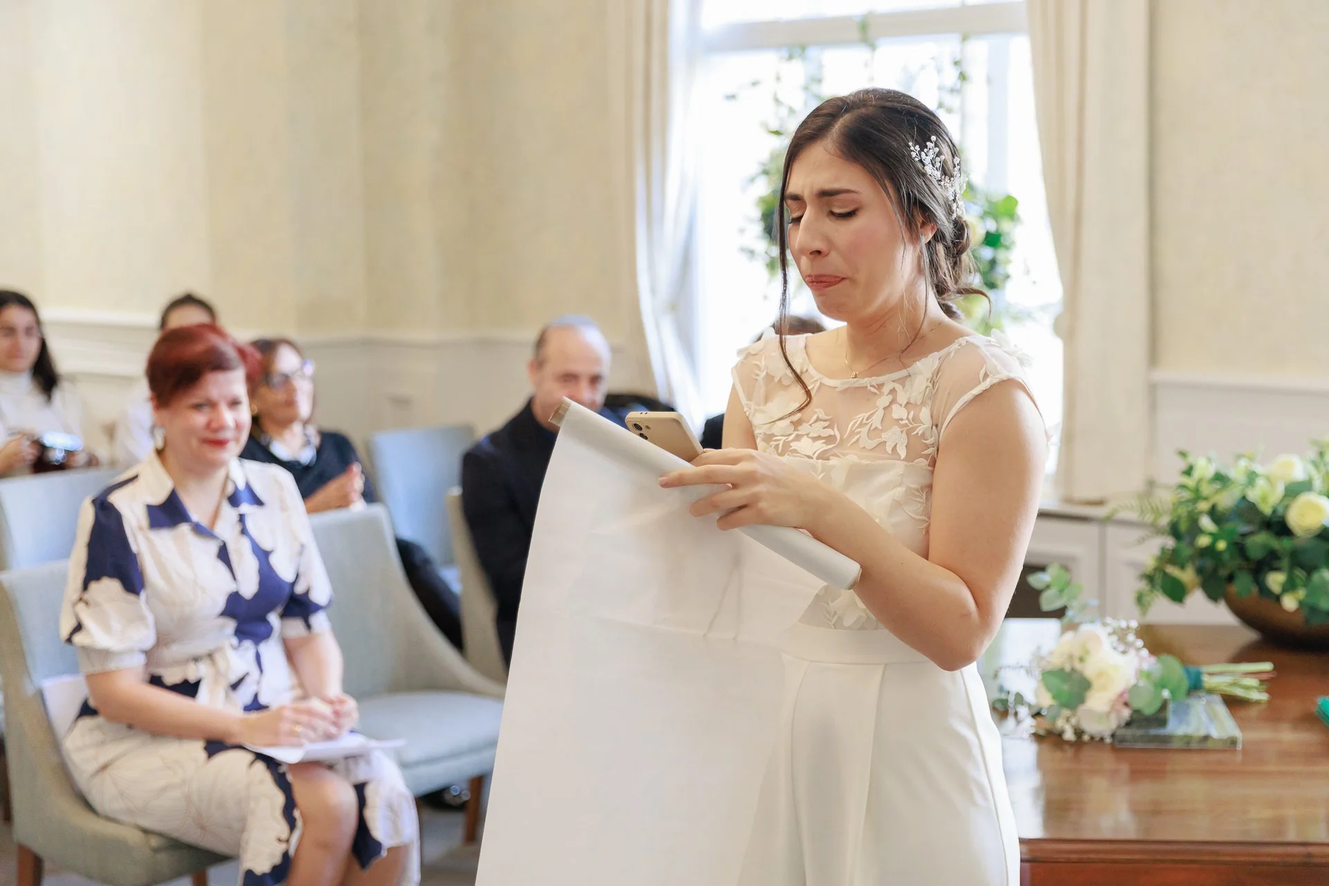 A woman in a white dress reading a note or card, appearing emotional, during a wedding ceremony with seated guests in the background.