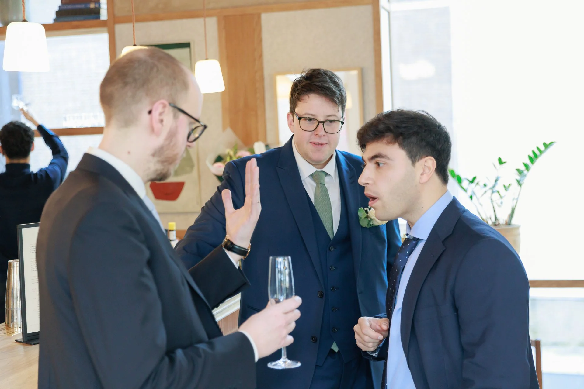 Three men in formal suits having a conversation at a social event, one holding a glass of champagne, with a woman in the background raising her arm.