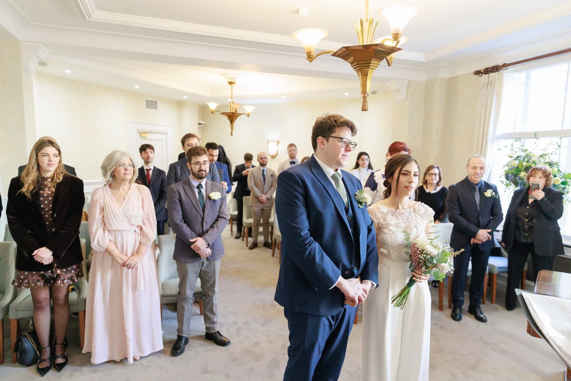 A wedding ceremony taking place in a bright room with large windows. The bride and groom are standing at the front, with the bride holding a bouquet. Guests are standing and observing the ceremony.
