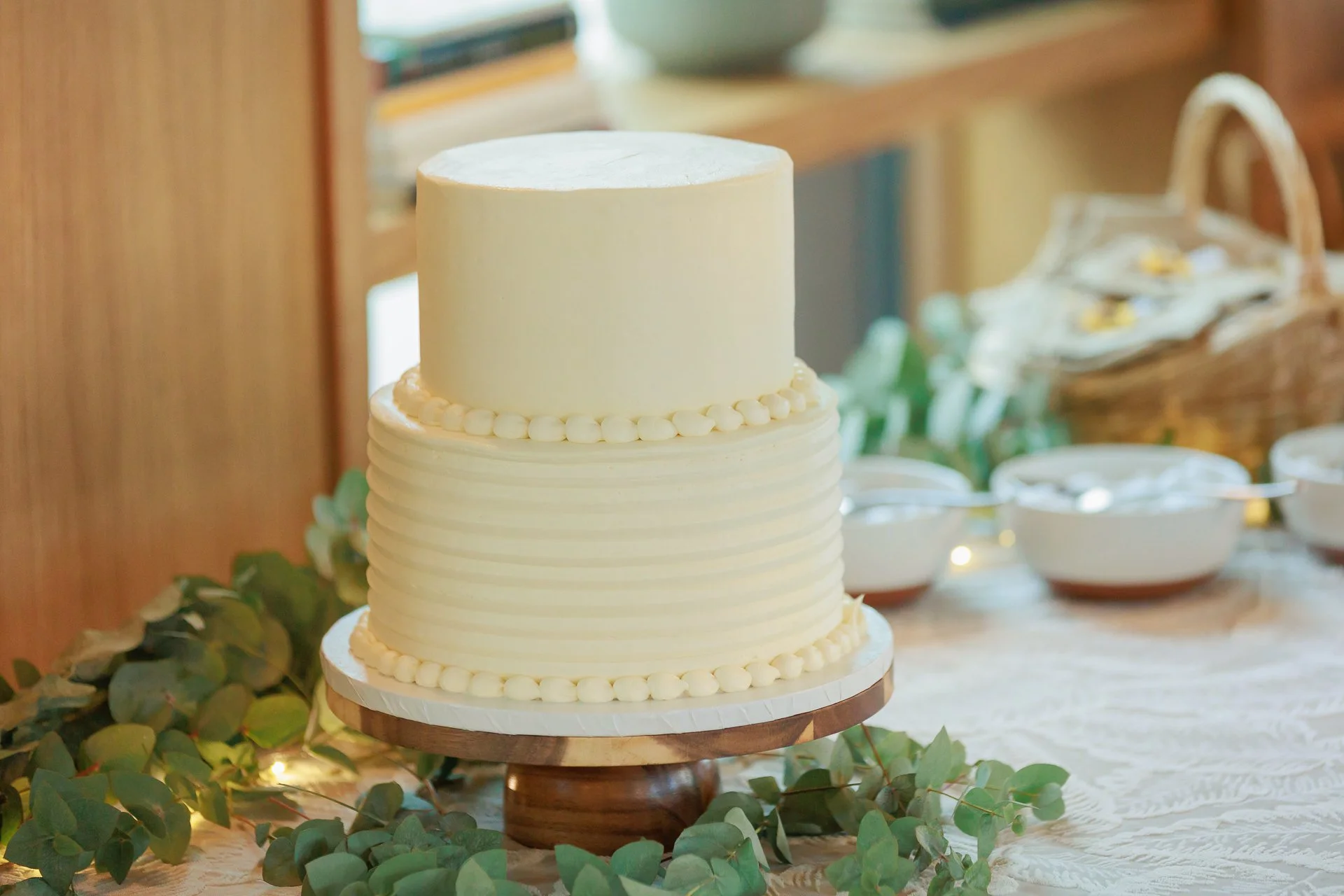 A two-tiered cream-colored wedding cake on a wooden cake stand decorated with small frosting pearls around the edges of each tier, placed on a table with greenery and bowls in the background.