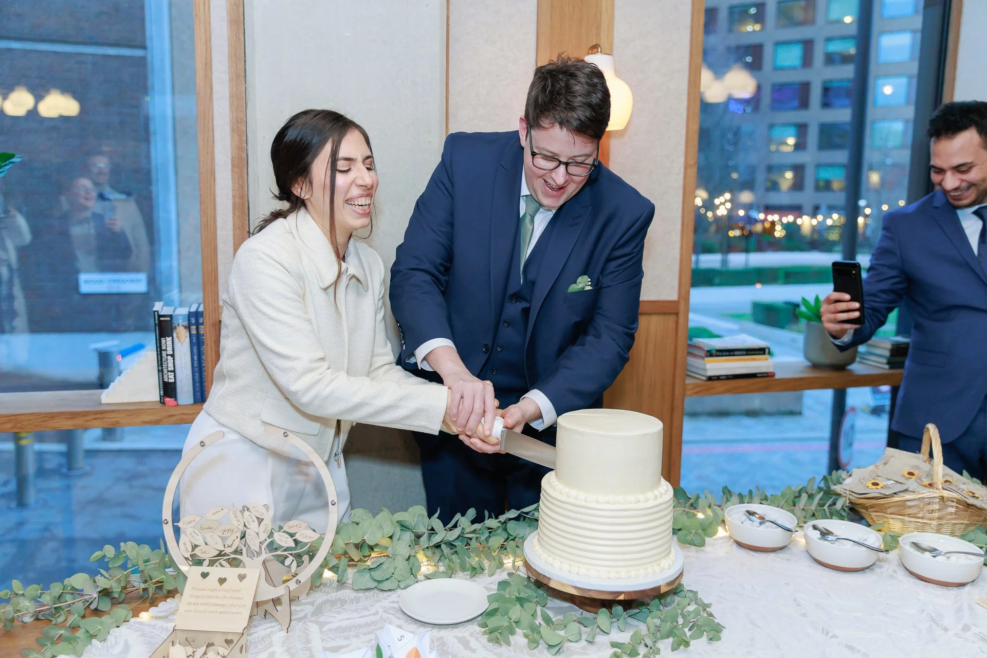 People celebrating and cutting a wedding cake at a reception, with a woman in a white coat, a man in a navy suit, and another man in a navy suit taking a photo. The table is decorated with greenery, bowls, and a small tree-shaped decoration.