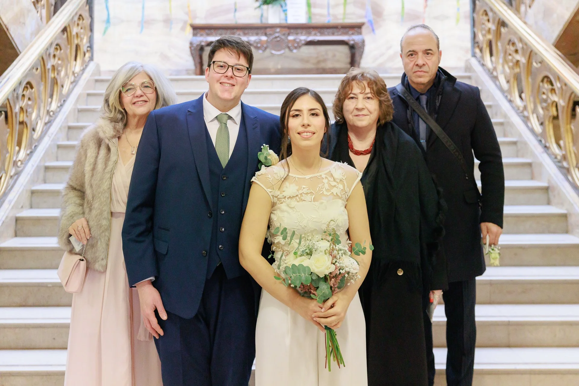 A group of six people, including a bride holding a bouquet, standing on a staircase inside a building with ornate gold railings, celebrating at a wedding.