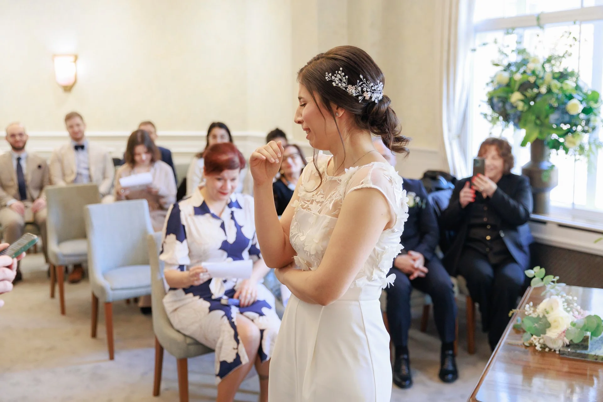 A bride in a white dress with floral details and a silver hairpiece standing in front of seated guests during a wedding ceremony or reception. Some guests are taking photos or looking at the bride, in a bright room with large windows and floral decor