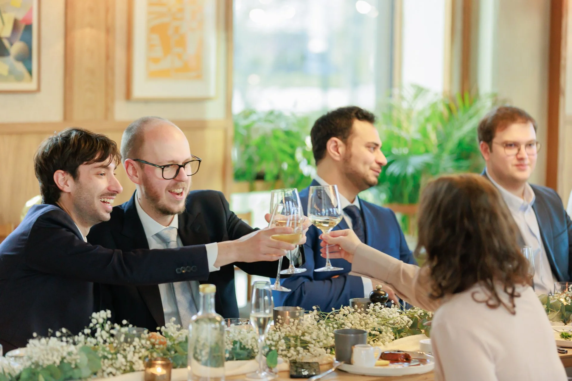 A group of five people dressed in formal attire, sitting at a decorated table, raising glasses for a toast during a celebration in a well-lit room with wooden paneling and green plants.