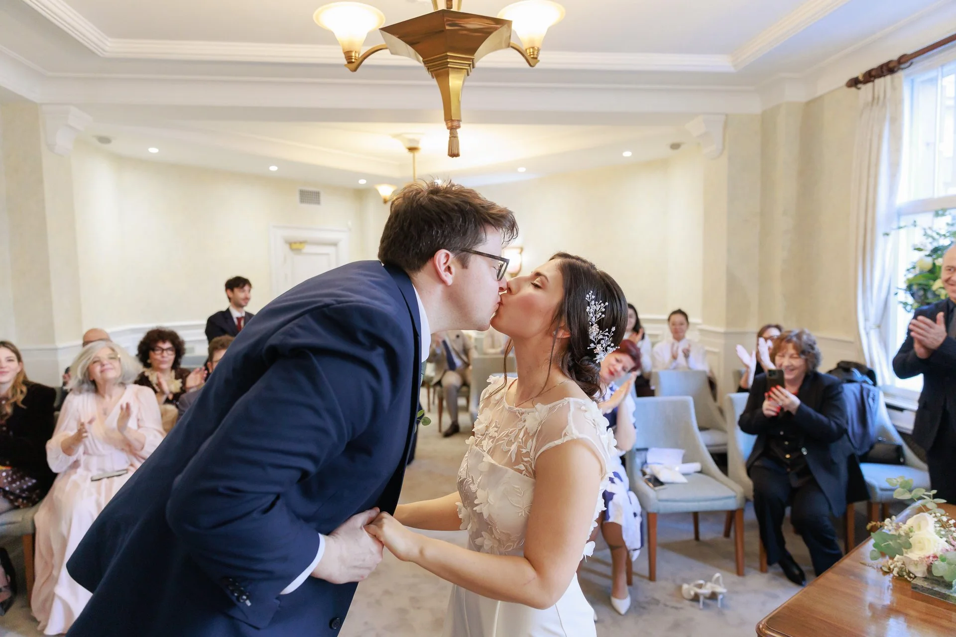 A newly married couple kissing during their wedding ceremony with guests clapping and smiling in the background.
