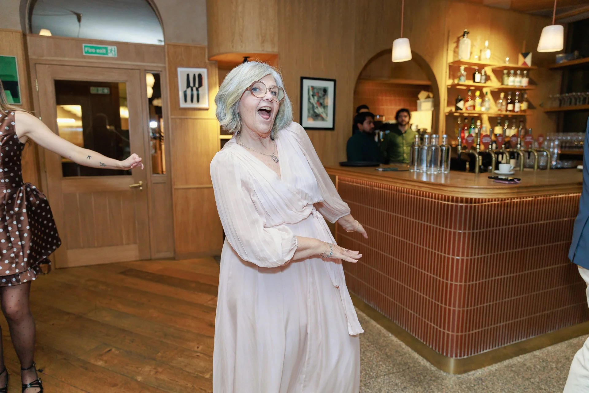 Older woman with light blonde hair, glasses, wearing a light pink dress, is dancing and smiling in a wooden-finished bar or restaurant, with bar staff and patrons in the background.