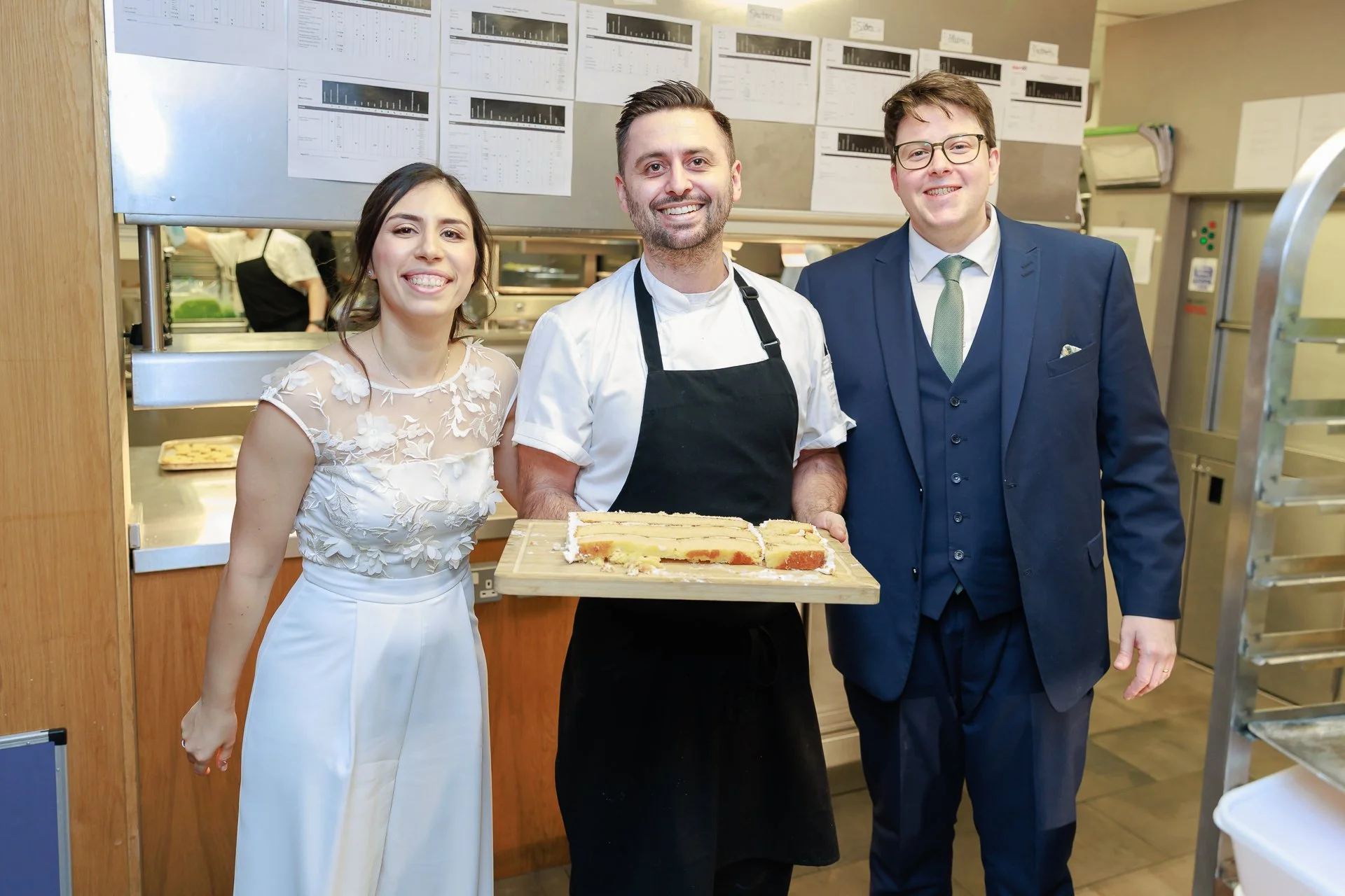 A woman in a white dress, a man in a white chef's coat and black apron, and a man in a blue suit with glasses standing together in a kitchen, with the chef holding sliced cake on a wooden board, all smiling at the camera.