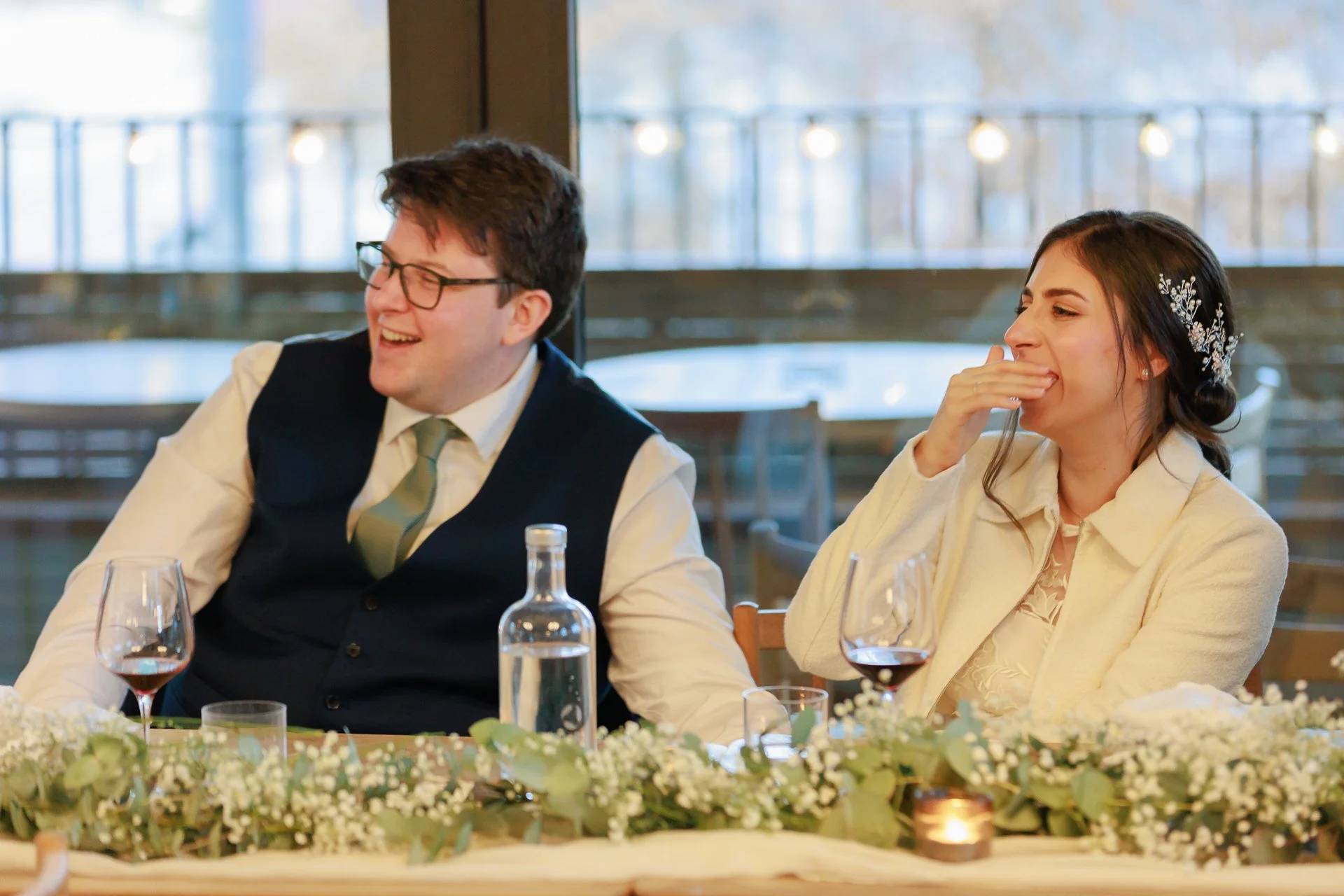 A man and woman sitting at a decorated table, laughing and enjoying a wedding reception, with wine glasses and a bottle on the table.