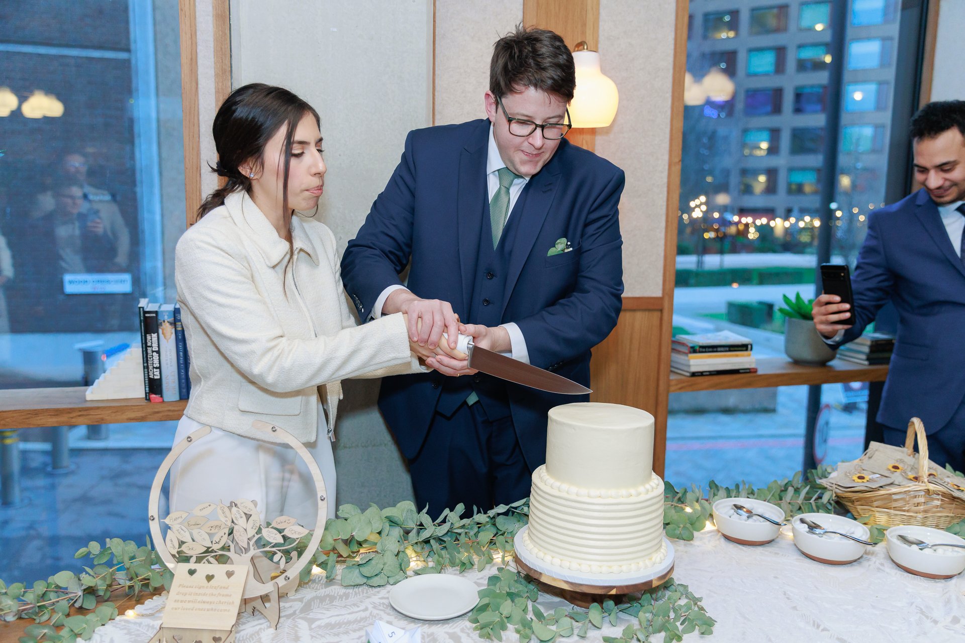 Two people, a woman and a man in formal attire, are cutting a wedding cake together at a reception. A third person is taking a photo in the background.