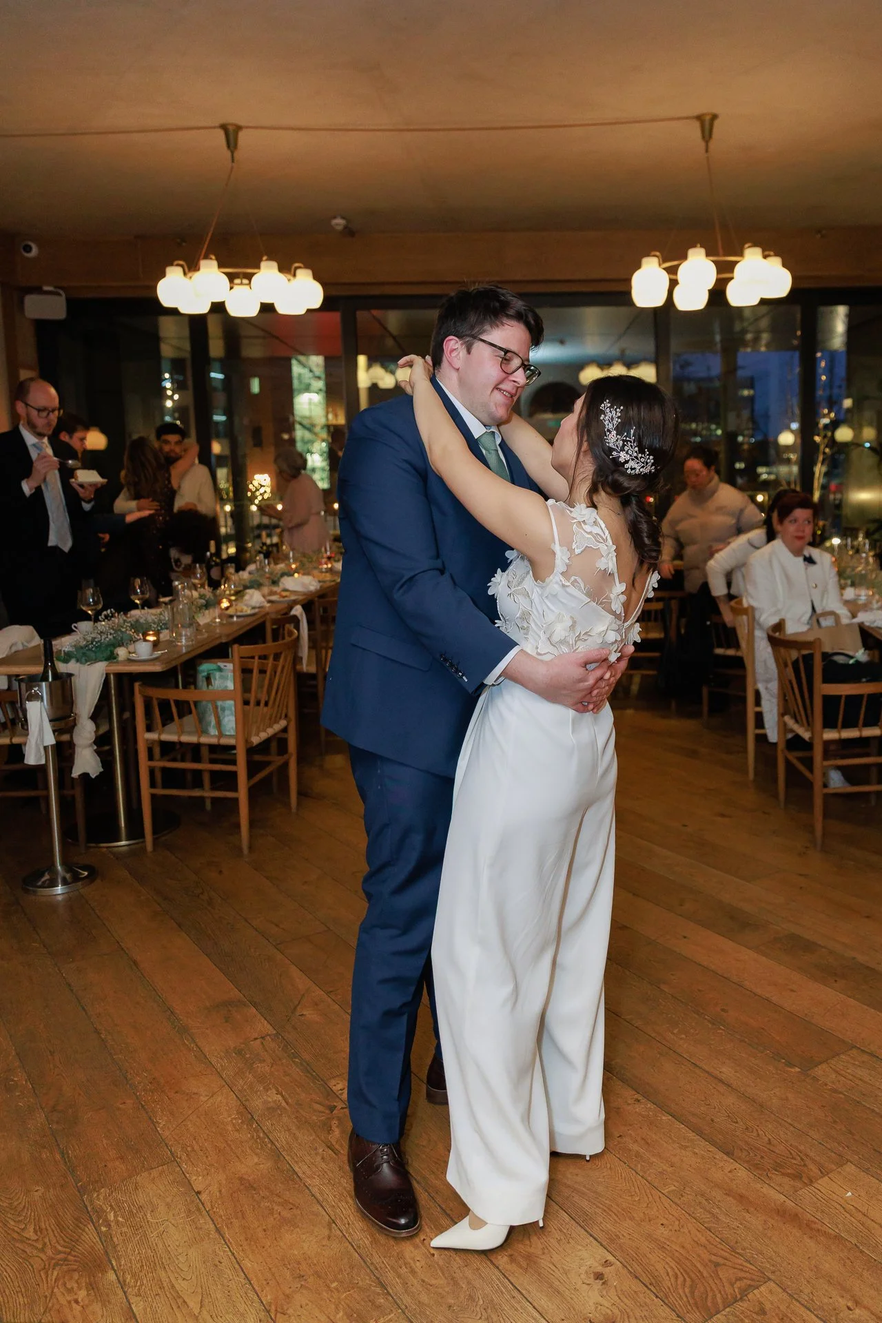 A bride and groom dancing at their wedding reception, holding each other closely and smiling, in a warmly lit room with guests seated at tables in the background.