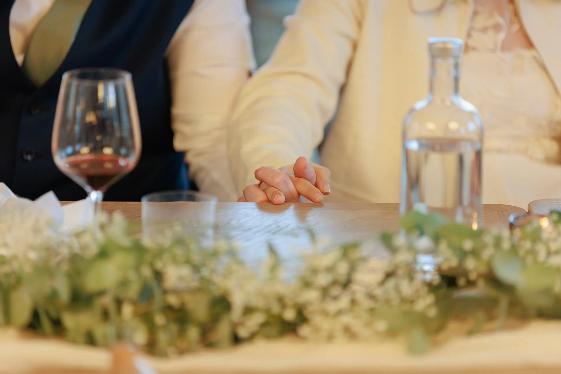 Close-up of a person with clasped hands resting on a table, with a glass of red wine, a water carafe, and a floral centerpiece in the foreground.