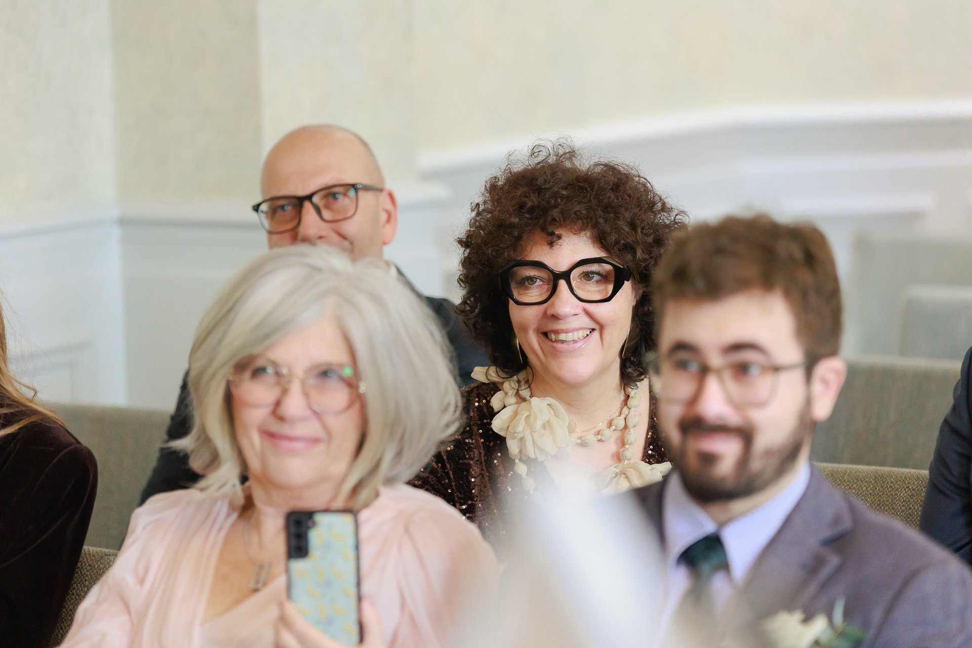 Group of people at a formal event, with three women and two men, smiling and looking towards the camera or a speaker, dressed in business or cocktail attire.