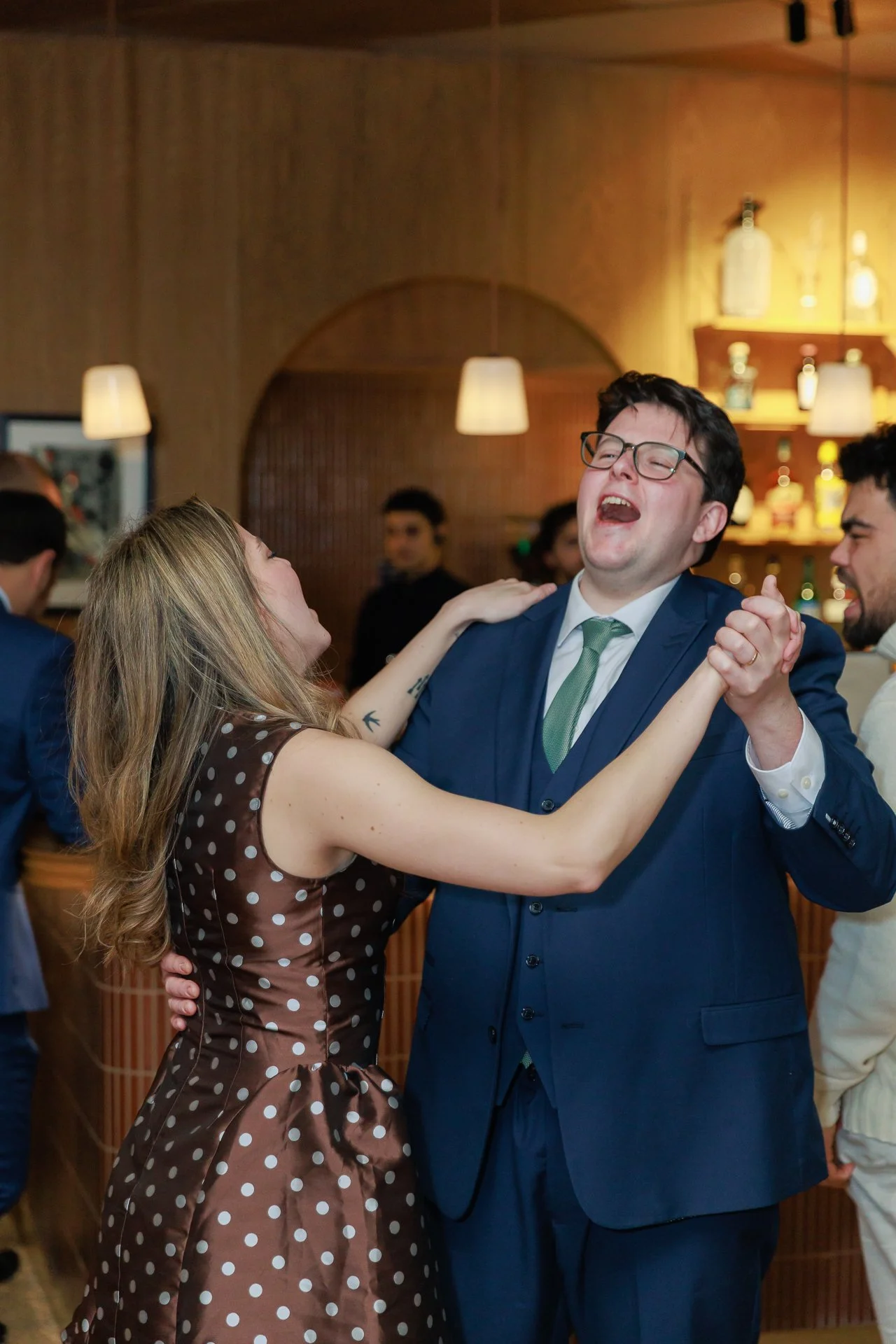 A woman and a man are dancing and laughing together at a party or celebration in a warmly lit room with other people in the background.