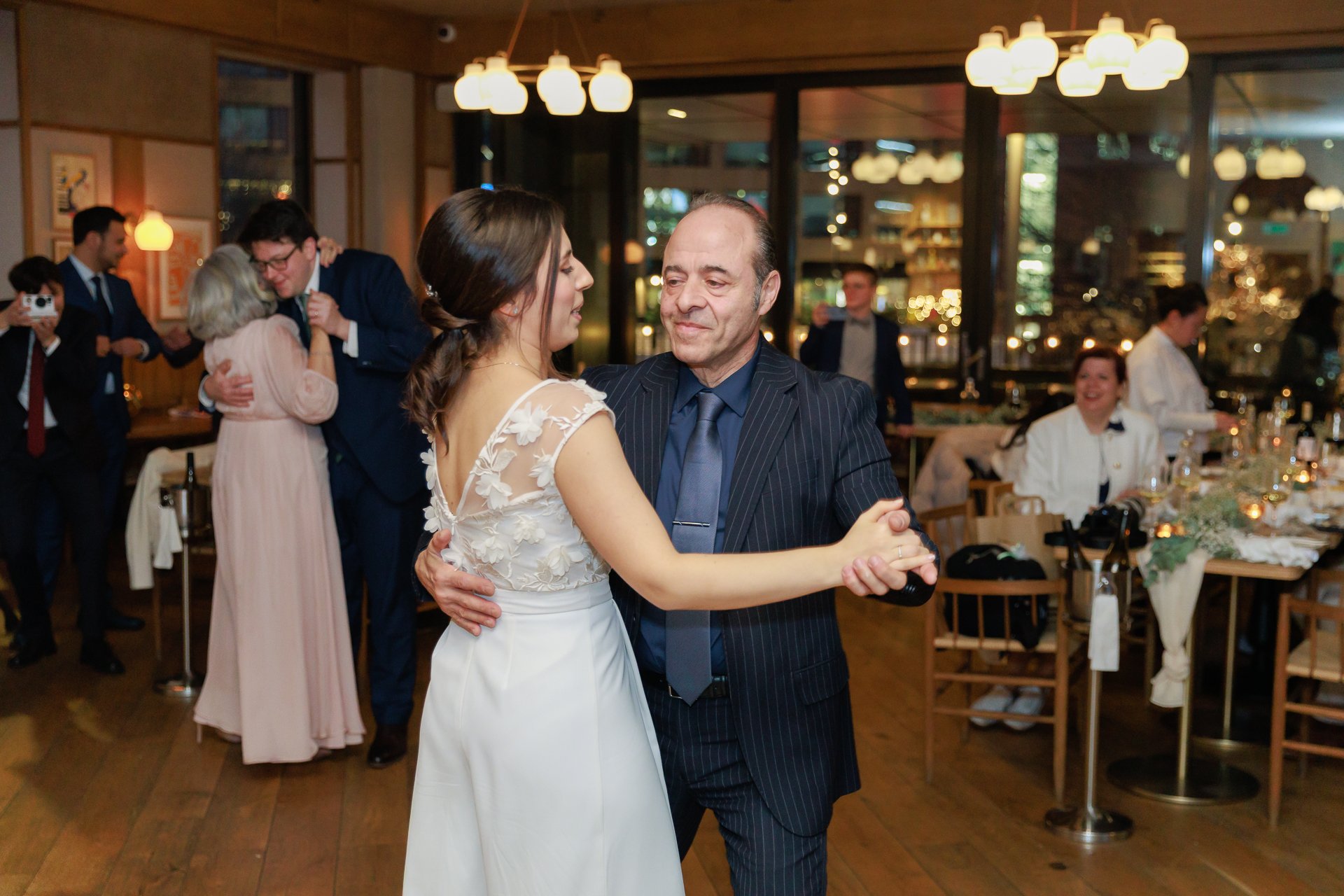 A bride and an older man dancing together at a wedding reception indoors, with other guests dancing and sitting at tables in the background.