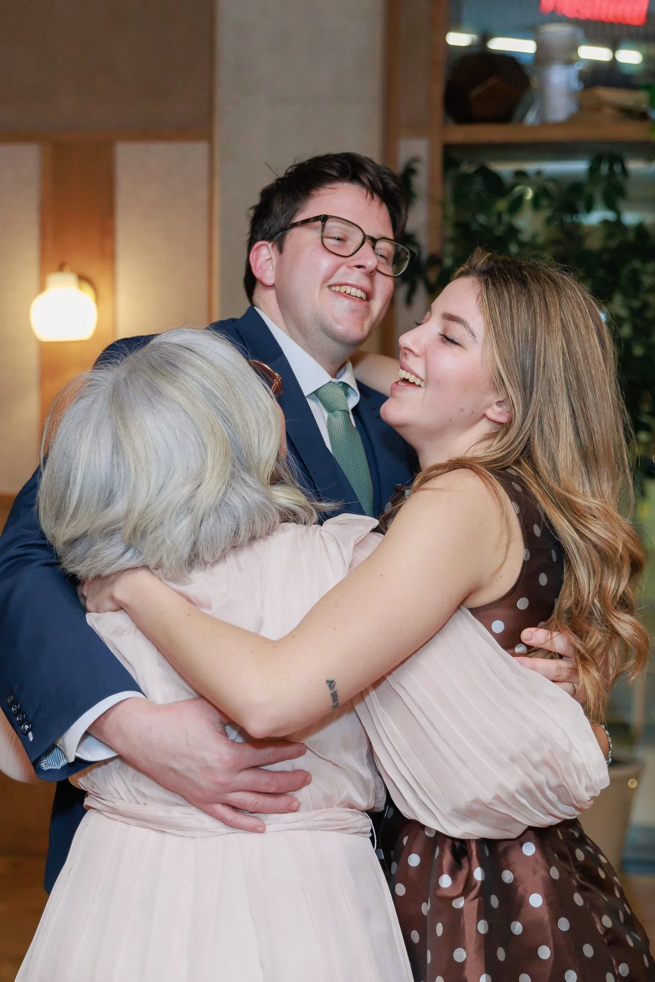 A man and woman are hugging, smiling, with a woman with gray hair also hugging them, in a warm indoor setting.