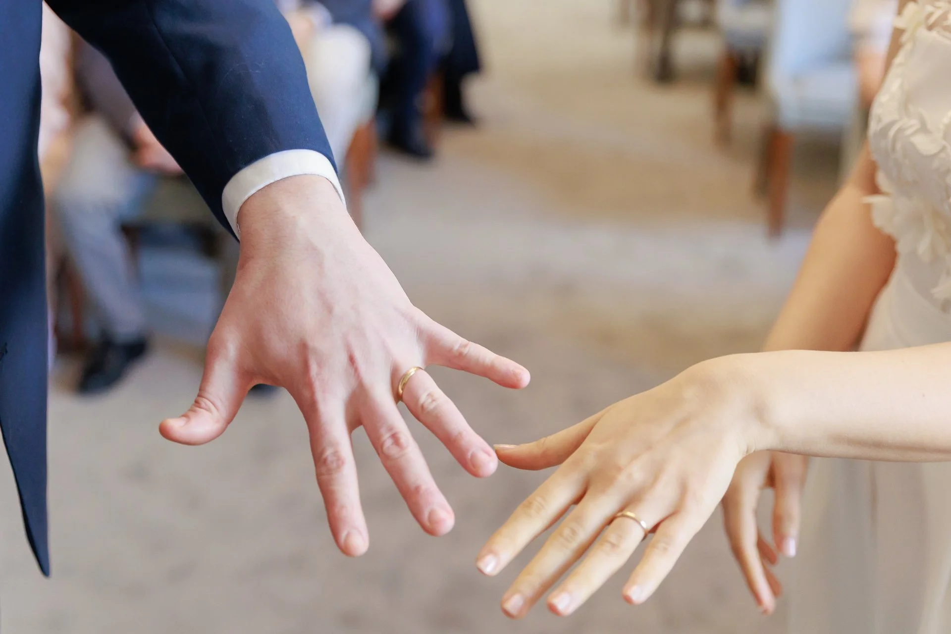 Close-up of a man and woman holding hands during a wedding ceremony, both wearing wedding rings, in an indoor setting with chairs and guests in the background.