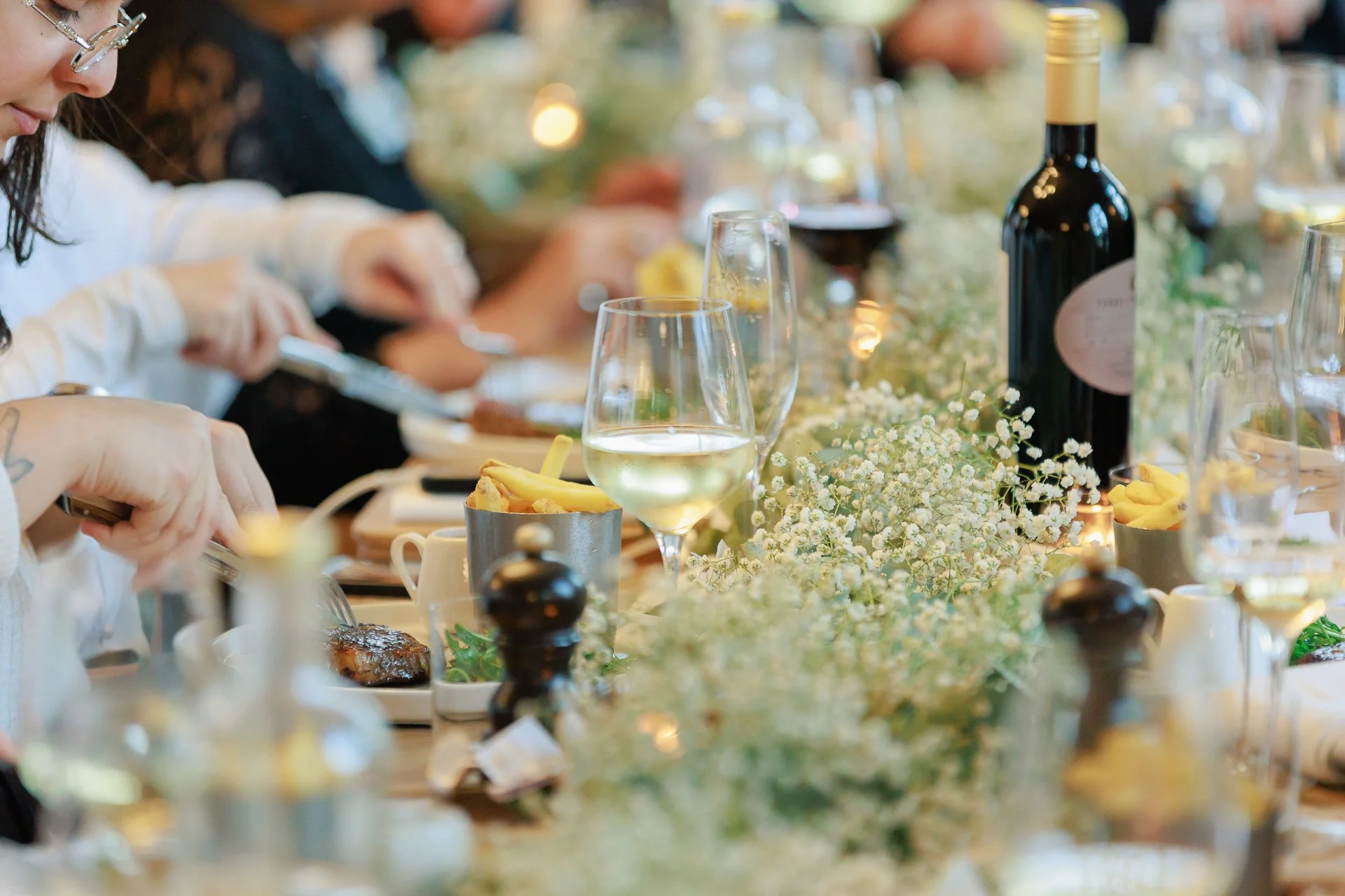 A close-up of a dining table set for a meal, with wine glasses, a bottle of wine, flowers, and plates of food, during a formal event.