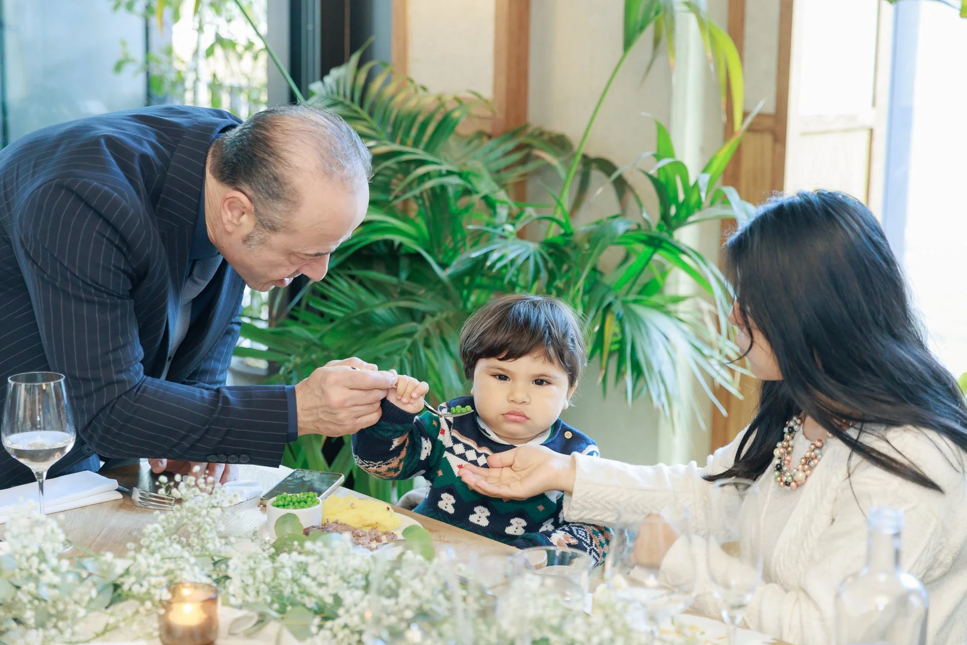 A young boy receiving food from an adult man at a family gathering, with a woman holding a spoon in front of him, in a room with green plants and natural light.