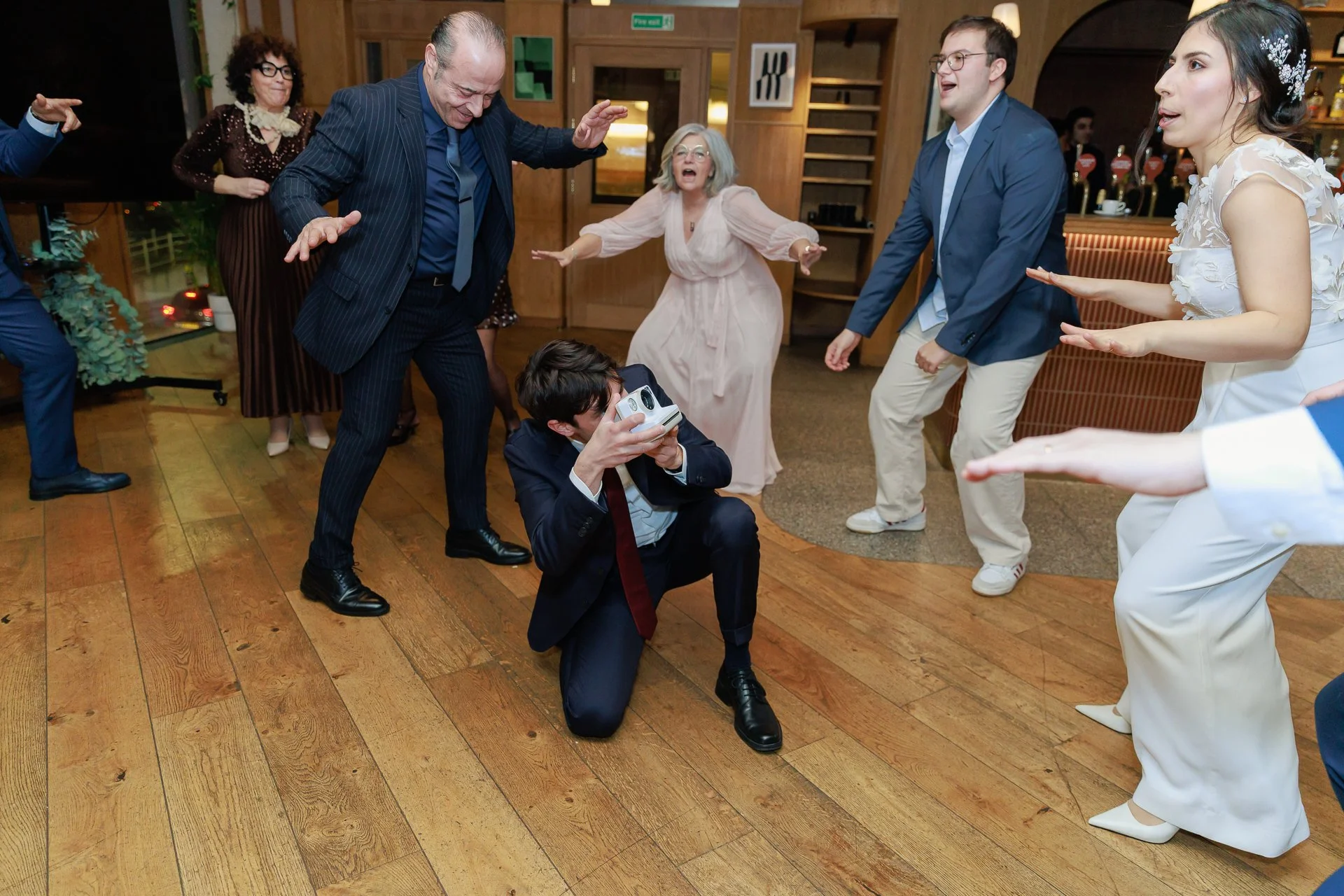 People dancing and celebrating at a party, with a man taking a photo or video using a vintage camera, in a lively indoor setting with wooden floors and warm lighting.