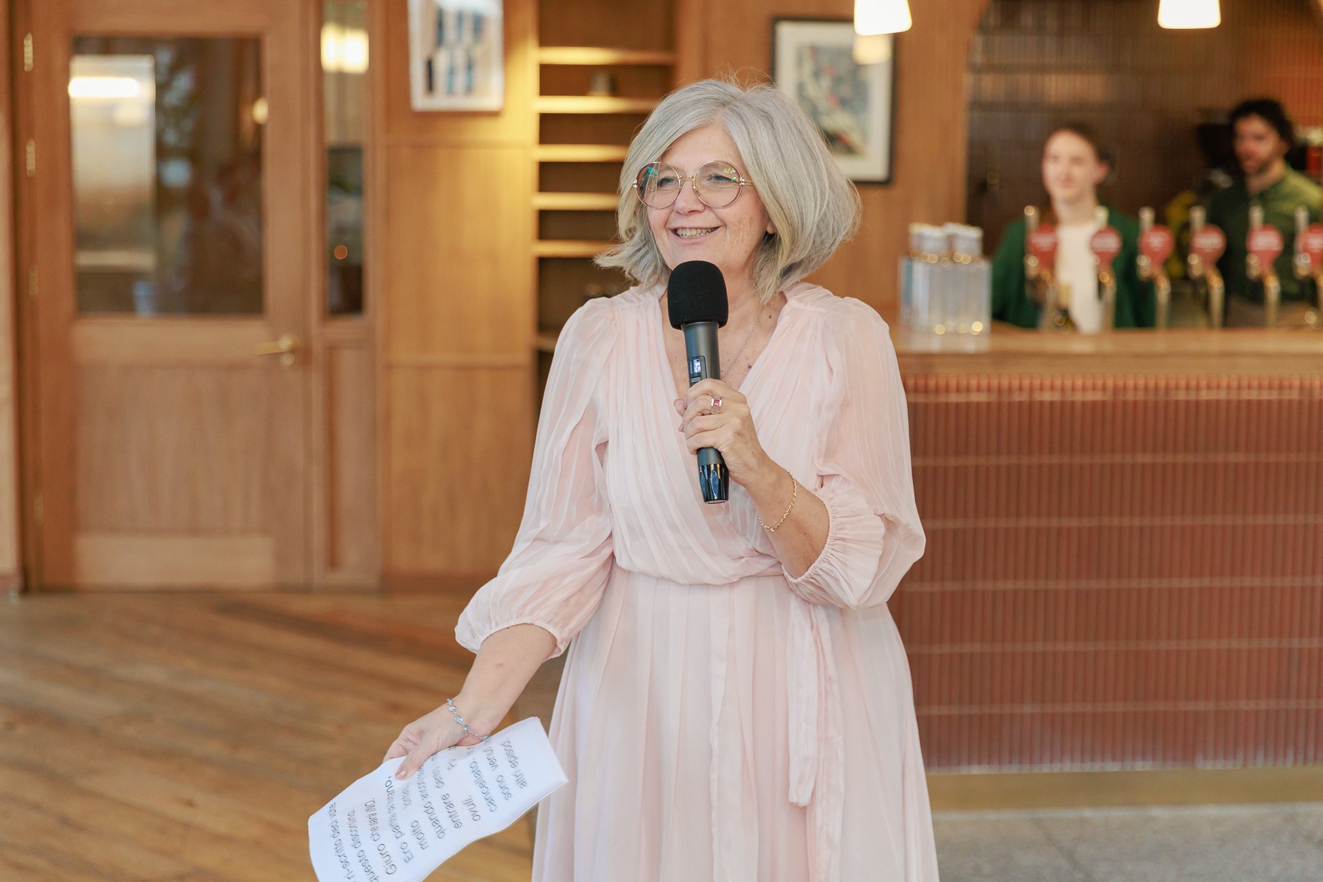A smiling older woman with gray hair and glasses holding a microphone and a piece of paper, standing indoors near a wooden bar with two women in green uniforms in the background