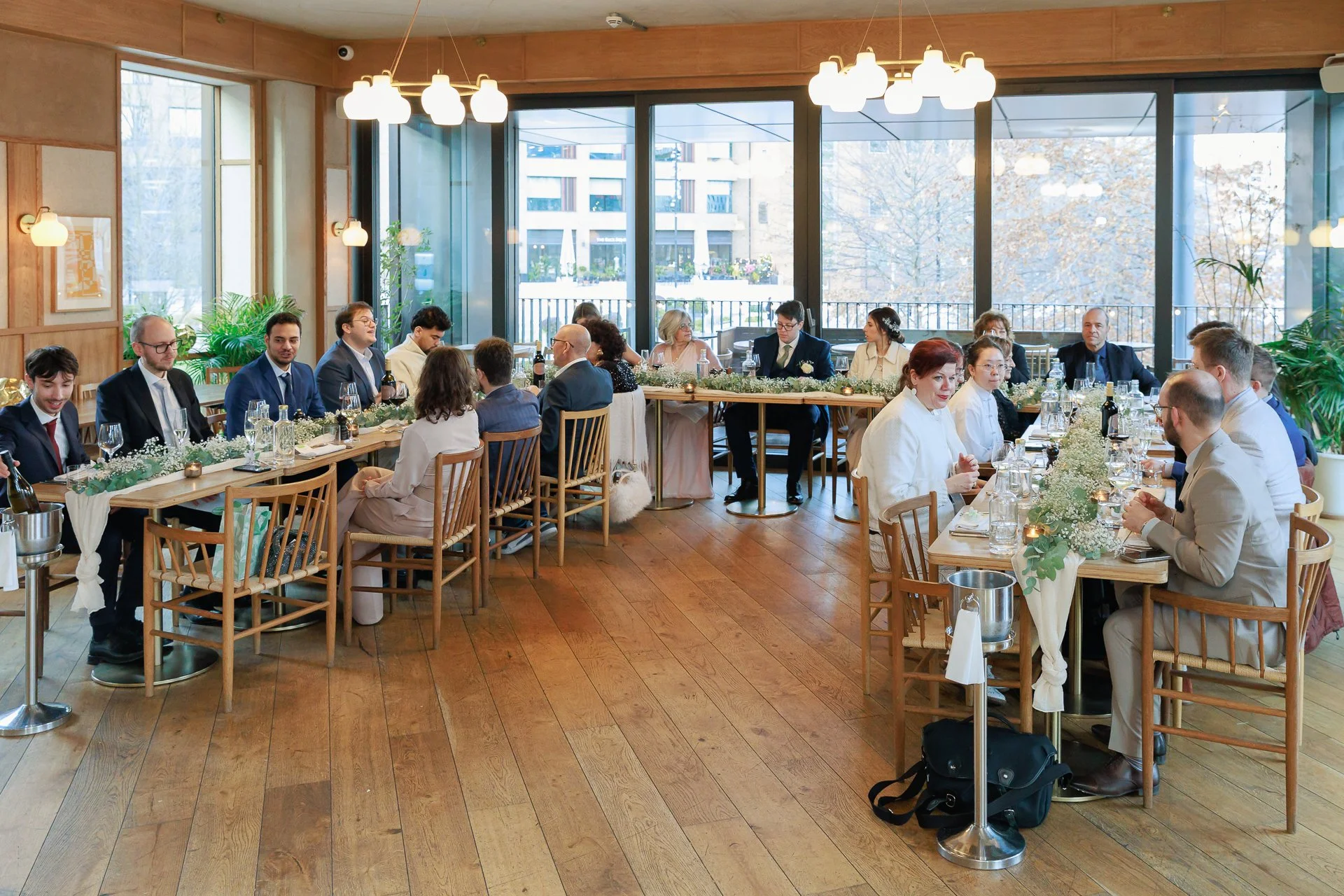 A formal wedding reception with guests seated at long rectangular tables decorated with flowers and candles, in a bright room with large windows and wooden floors.