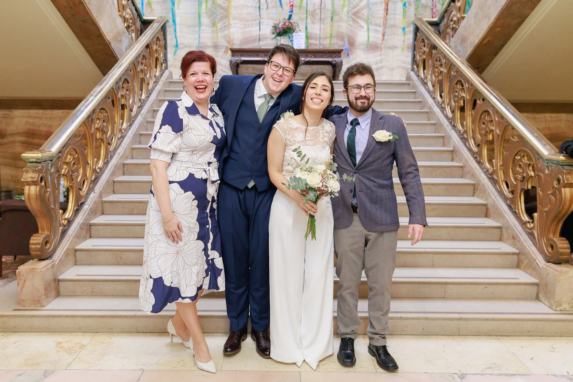 Group of four people, three men and one woman, smiling and posing on a staircase at a wedding celebration. The woman is holding a bouquet, wearing white, and the others are dressed in formal attire.