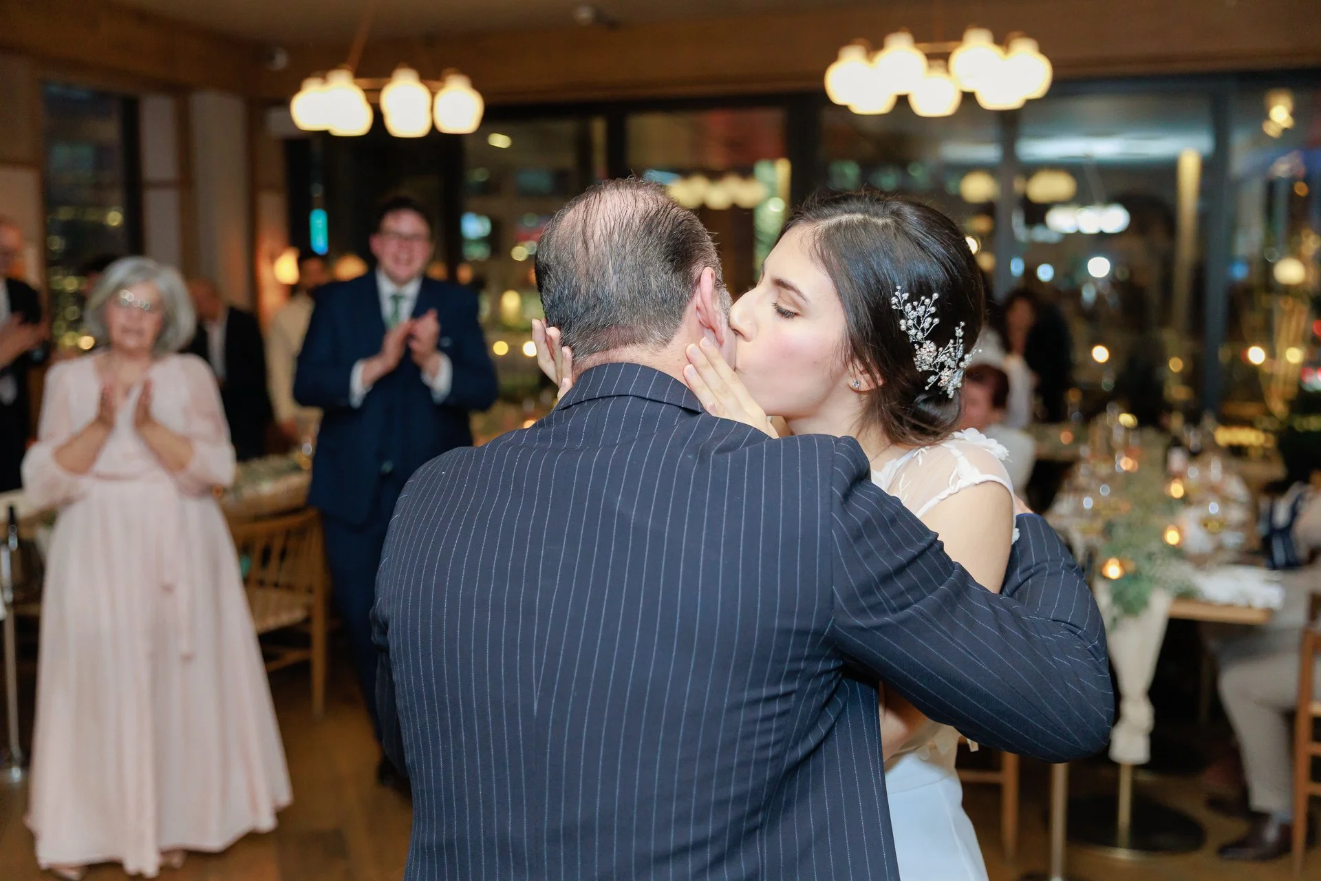 A bride and groom share a kiss during their wedding reception, surrounded by family and friends in a warmly lit venue.