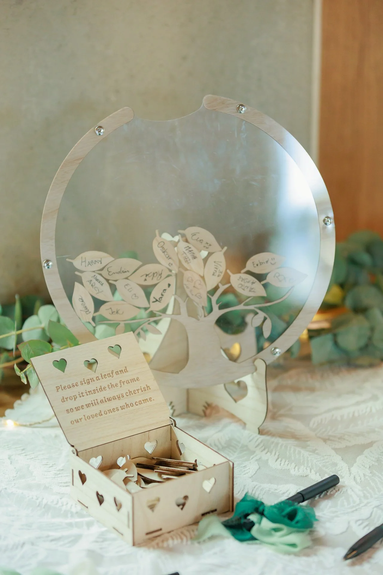 A decorative wooden family tree frame with paper leaves, each containing a name, displayed on a table with an open box of paper leaves, pens, and a note instructing guests to sign a leaf.