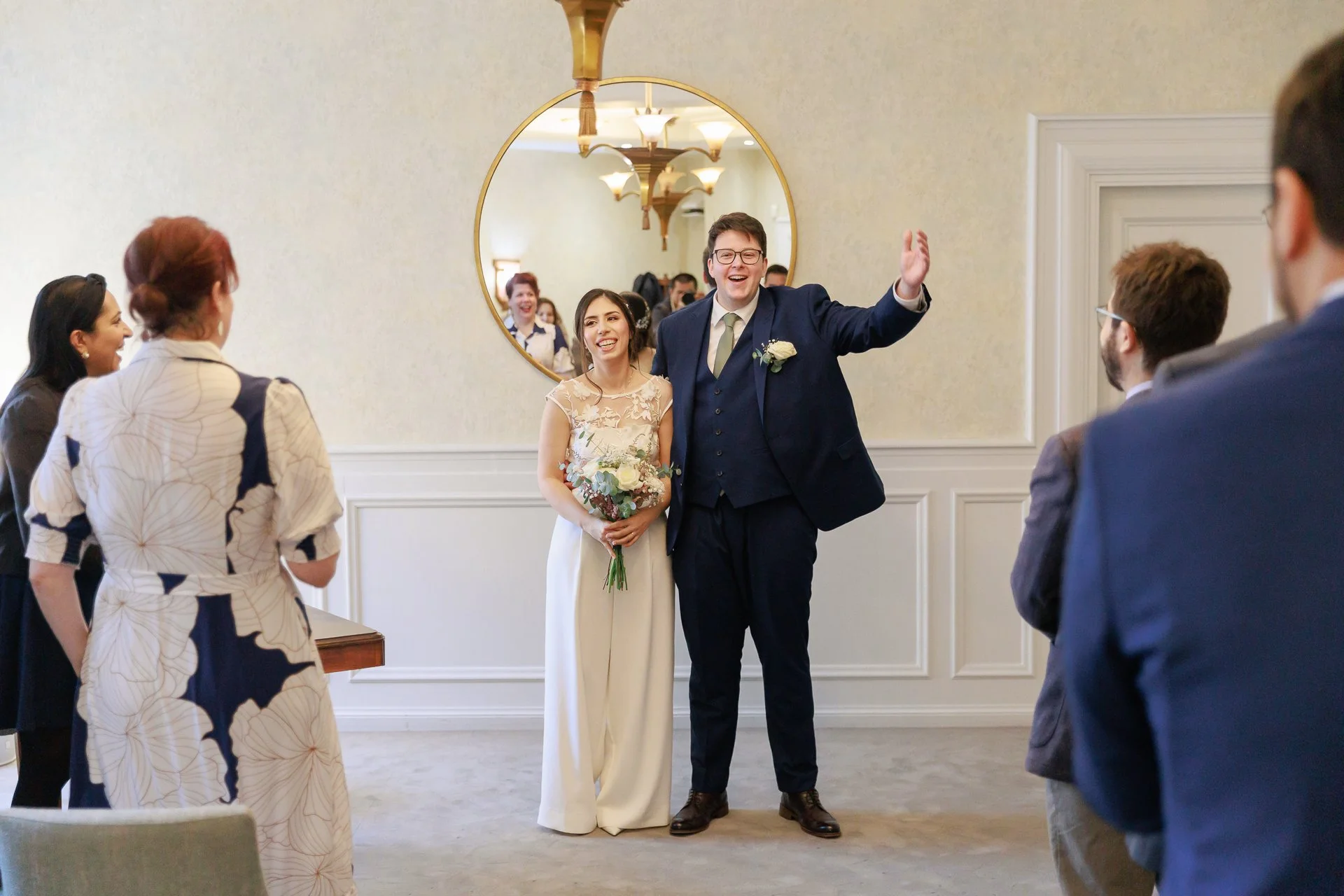 A wedding reception with the bride and groom standing in front of a mirror, surrounded by friends and family. The groom is wearing a navy suit and glasses, and the bride is holding a bouquet. The mood is joyful and celebratory.