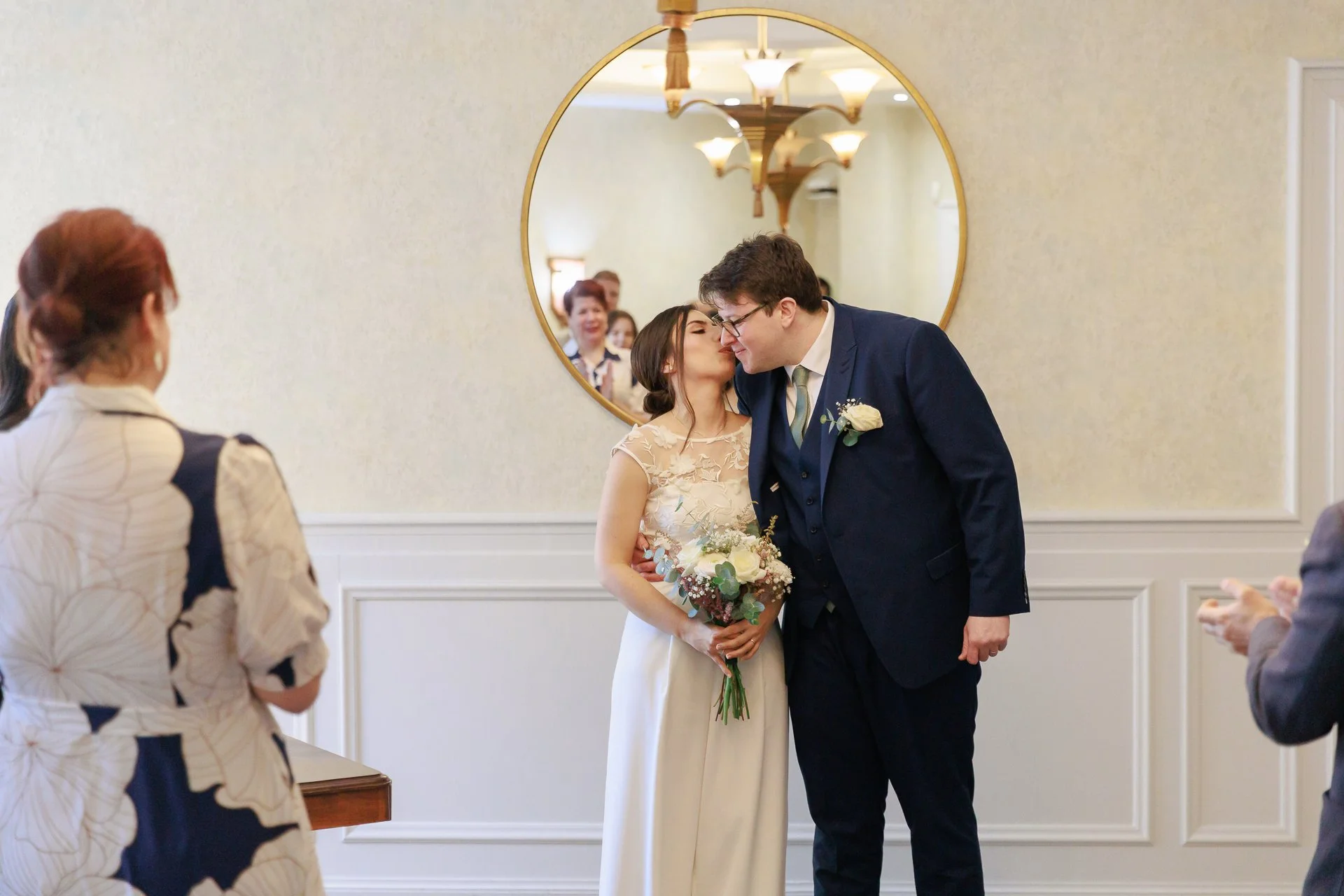 A wedding ceremony with a bride and groom kissing, surrounded by guests, in a room with a mirror and chandelier reflections.