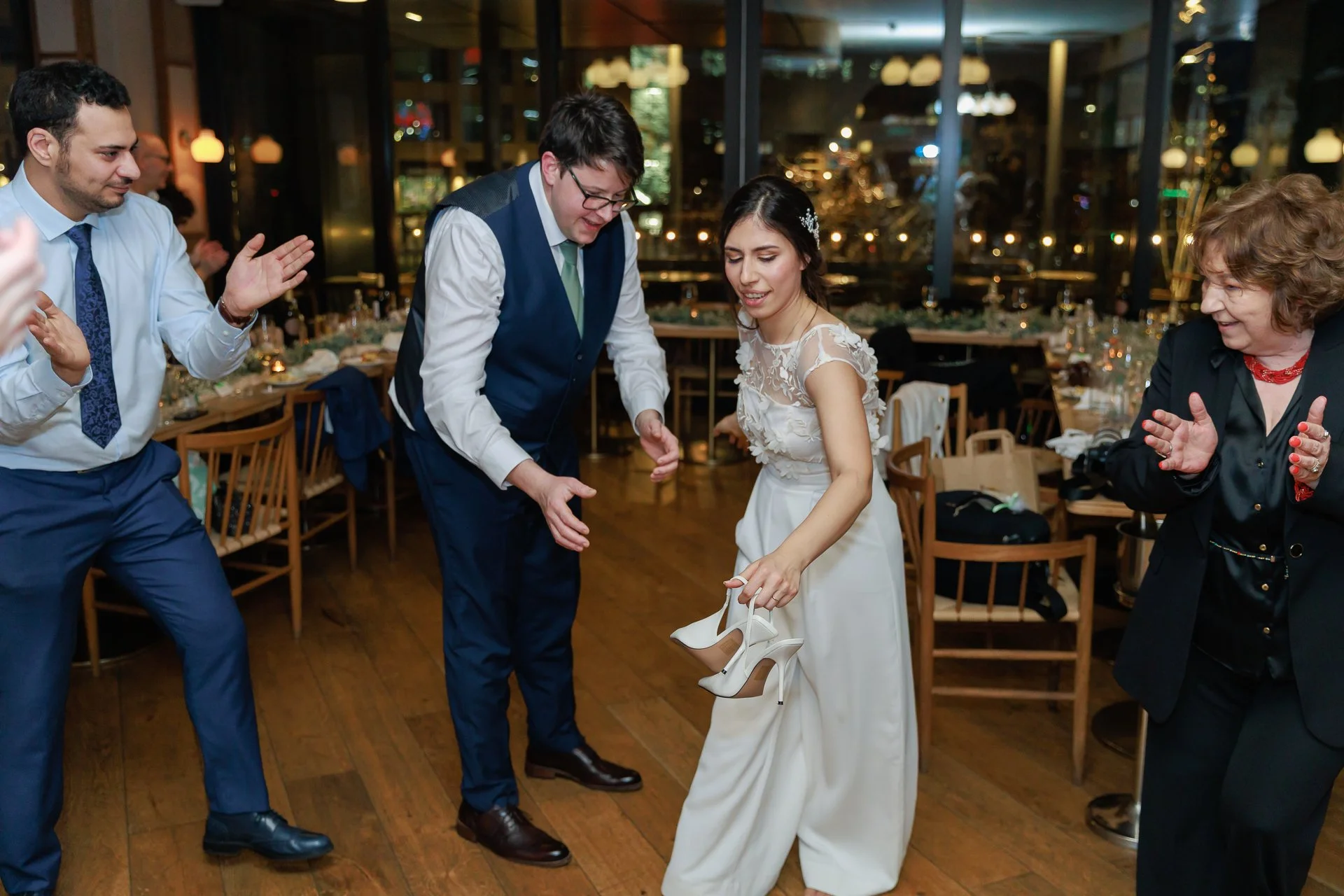 Group of people dancing at a wedding reception, with a woman in a white wedding dress holding high-heeled shoes, surrounded by others clapping and celebrating in a decorated venue with large windows and ambient lighting.
