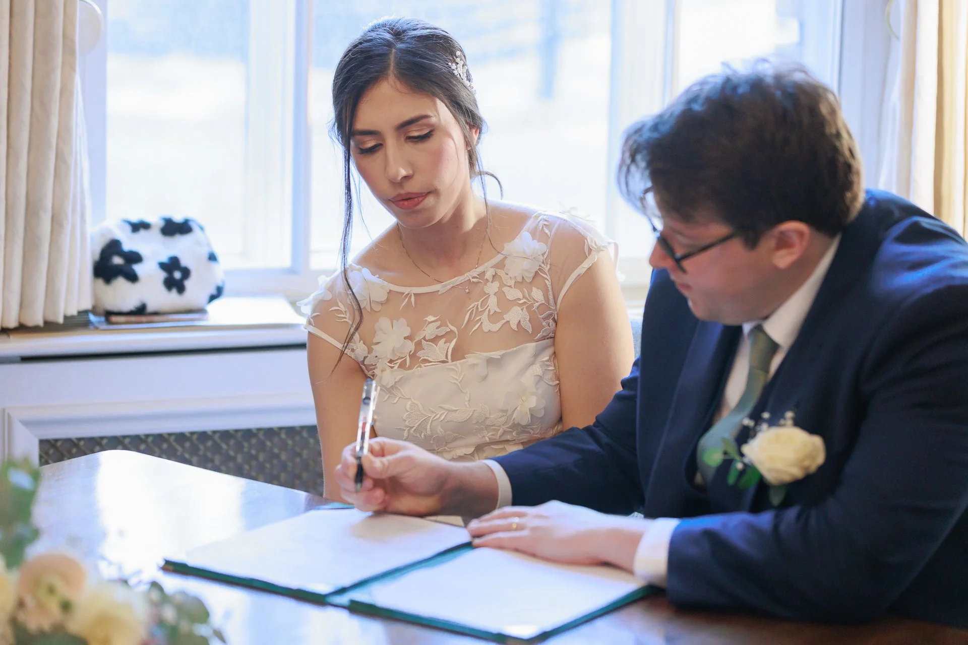 Bride in a wedding dress signing a document at a wedding ceremony while groom in a suit watches.