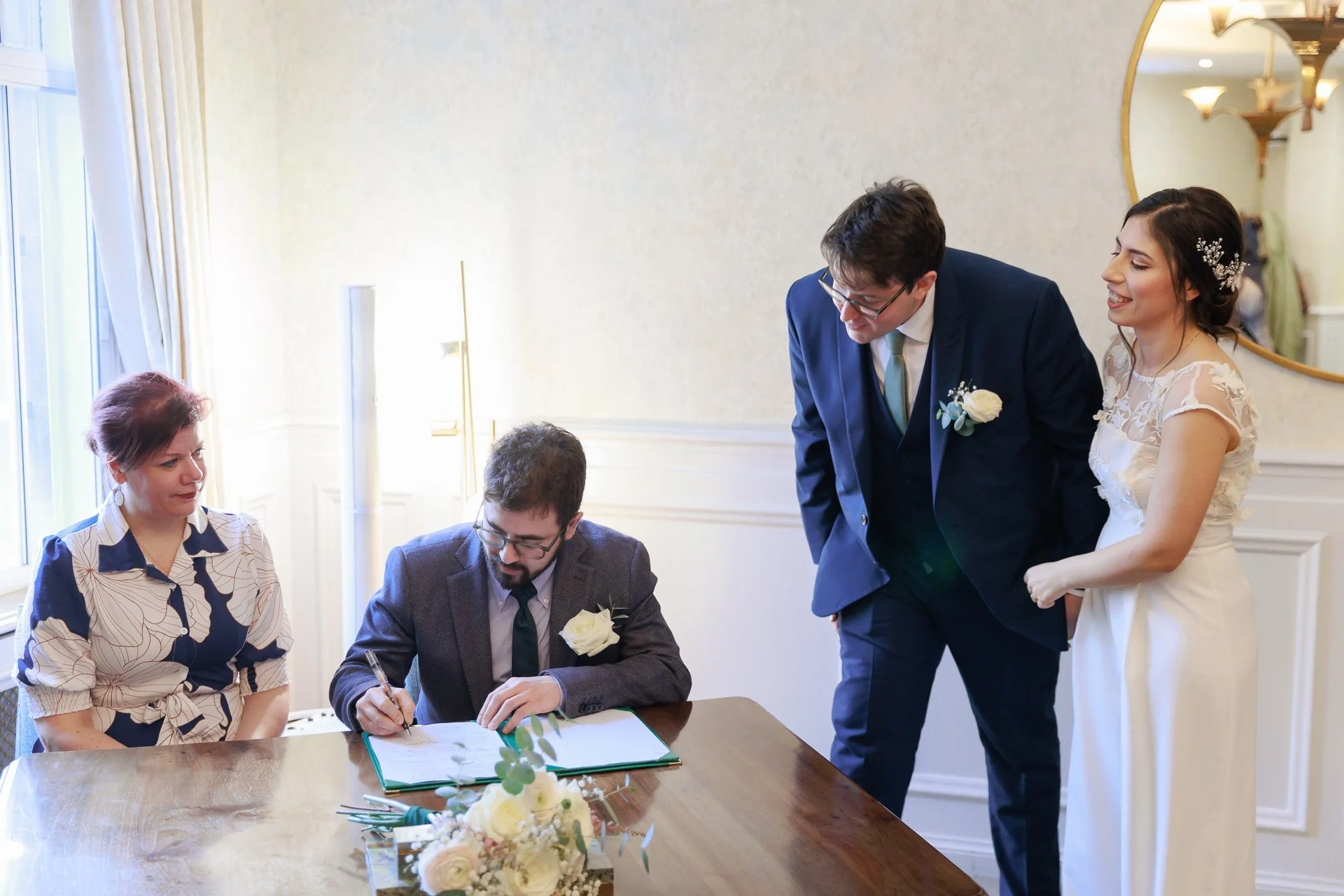 A wedding ceremony with a bride and groom signing a document at a table, guests seated nearby, flowers on the table, in a bright room with a mirror on the wall.