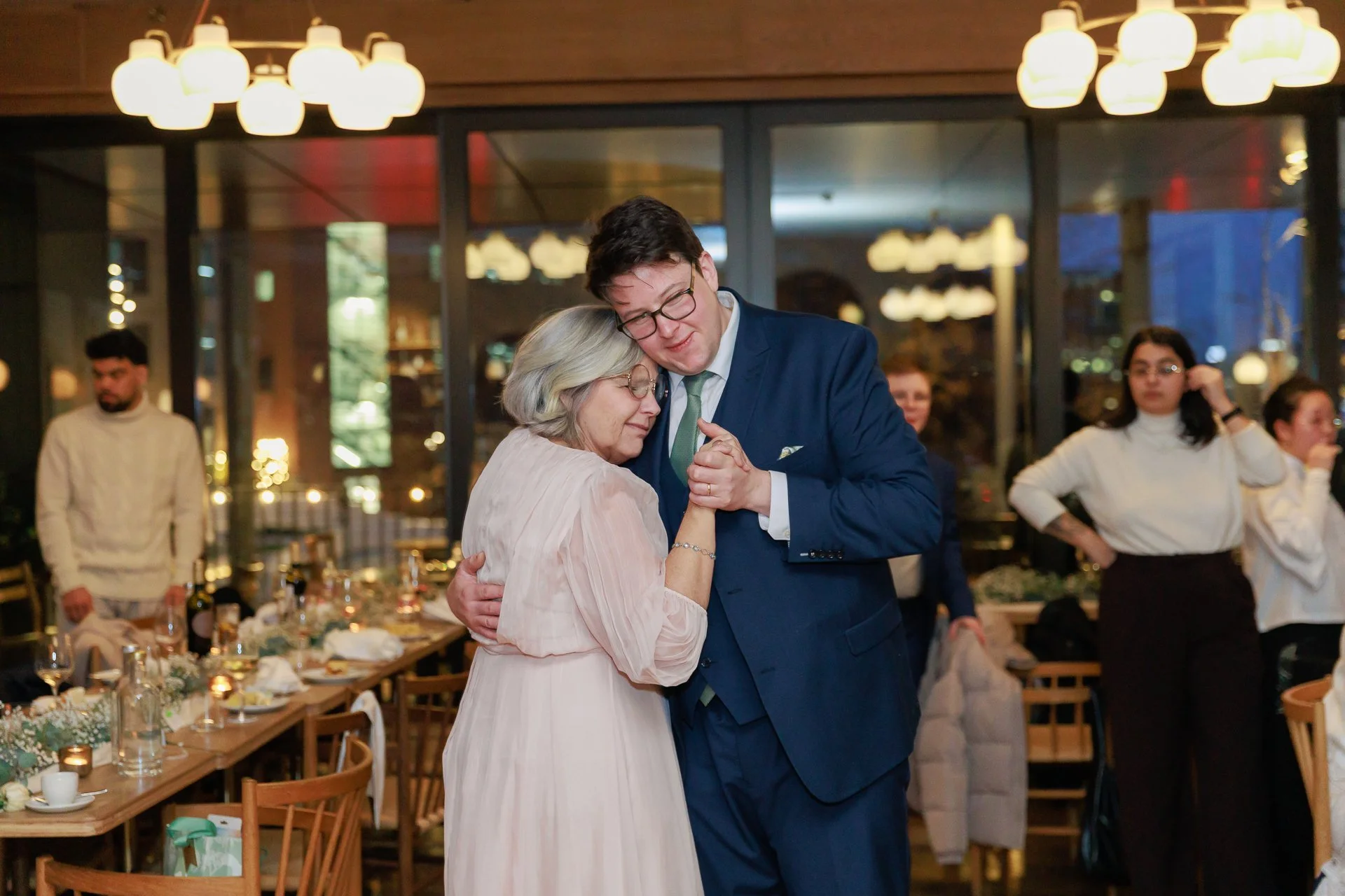 A man and woman dancing closely at a celebration or gathering, with people in the background in a warmly lit room with large windows and framed by hanging lights.