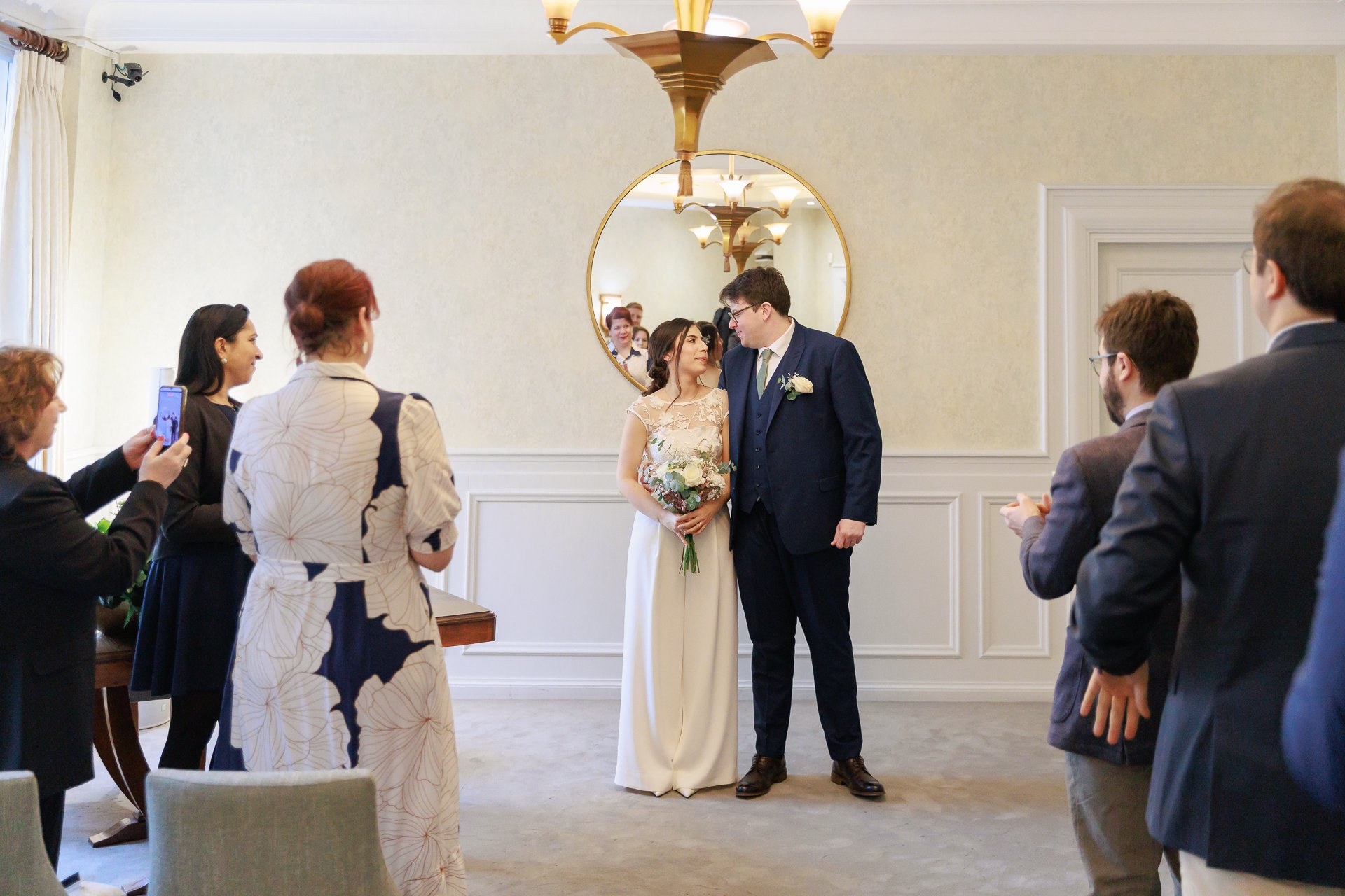 Bride and groom standing together, smiling at each other during their wedding ceremony, surrounded by friends and family taking photos, in an elegant room with a large round mirror and chandelier.