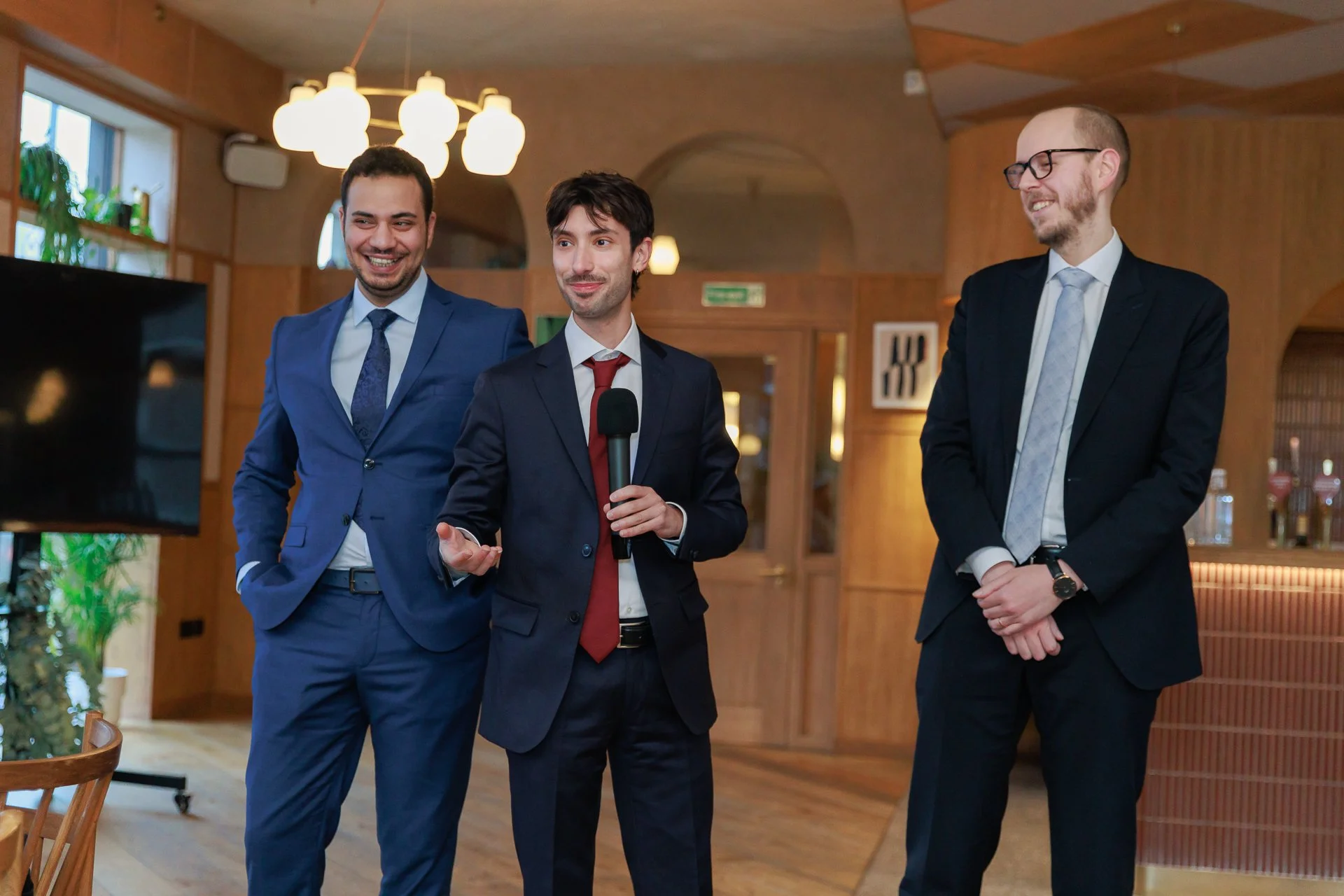 Three men in suits standing in a room, one holding a microphone, smiling, and appearing to give a speech or presentation.