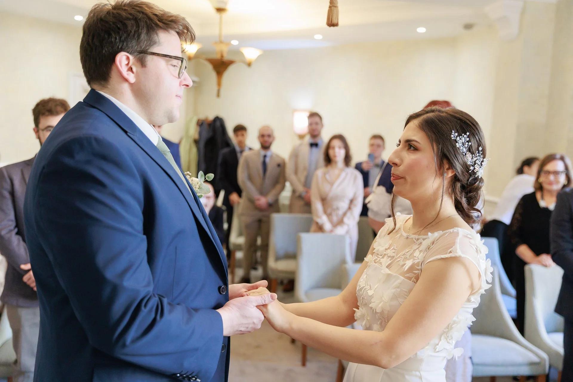 A bride and groom holding hands during their wedding ceremony, standing before family and friends in a decorated indoor venue.