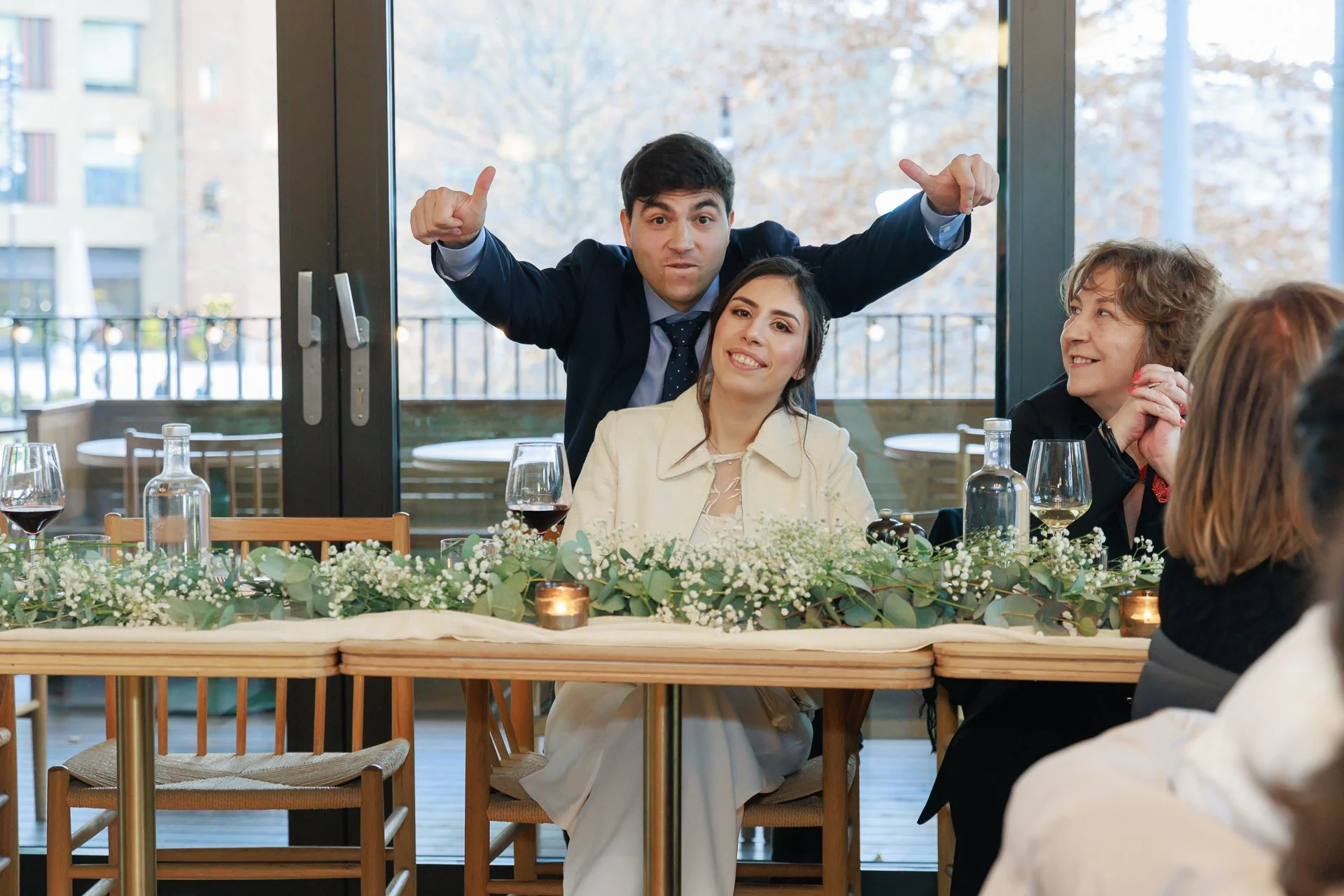 People celebrating at a formal event with a decorated table, wine glasses, bottles, and flowers, indoors near large windows.