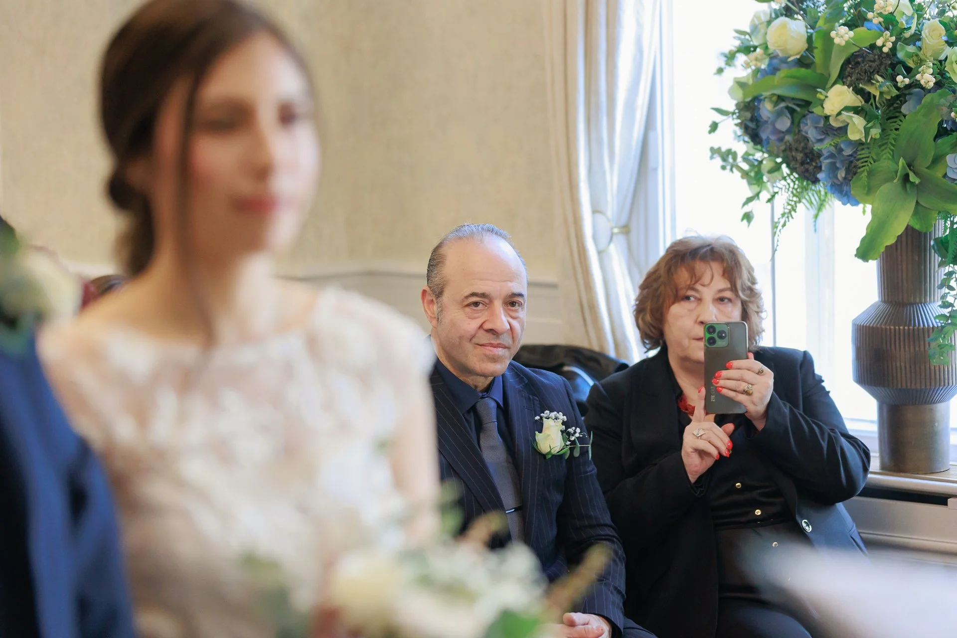 A wedding scene with a blurred bride in the foreground, a middle-aged man in a suit with a boutonniere looking at the camera, and a woman with curly hair taking a photo with her phone, inside a decorated room with flowers and a large window.