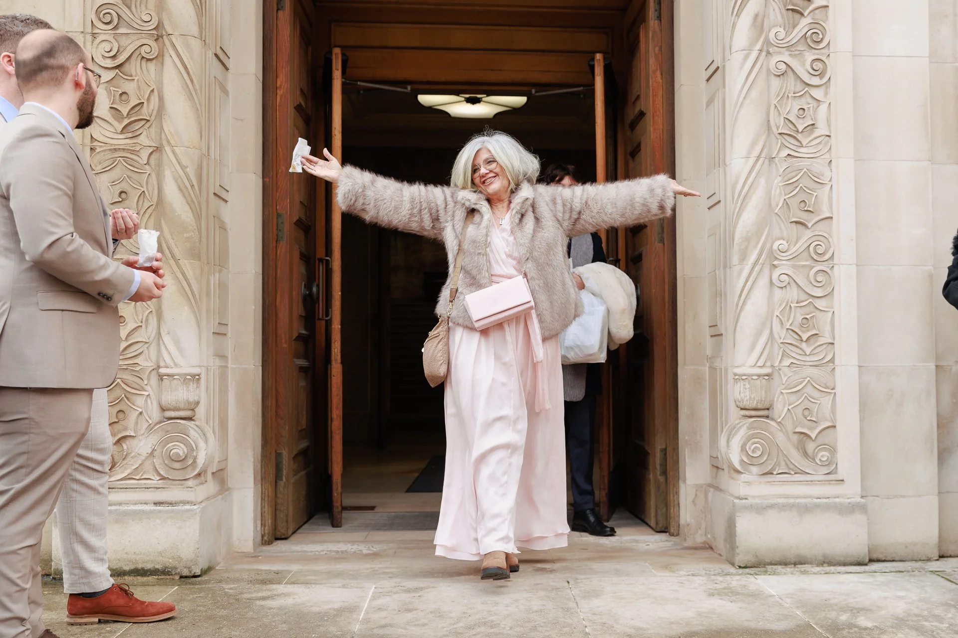 A joyful woman with gray hair and glasses, wearing a pink dress, a faux fur coat, and carrying a pink clutch, happily greets guests at the entrance of a building with ornate stone details, with arms wide open.