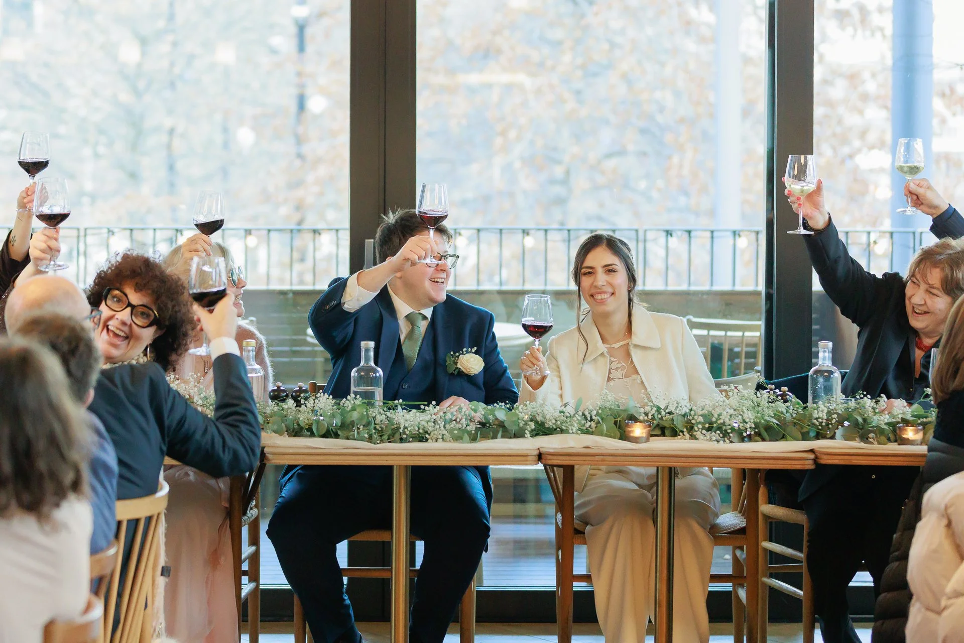 Wedding celebration with guests raising glasses of red and white wine, seated at a decorated table indoors with large windows and a view of autumn trees.