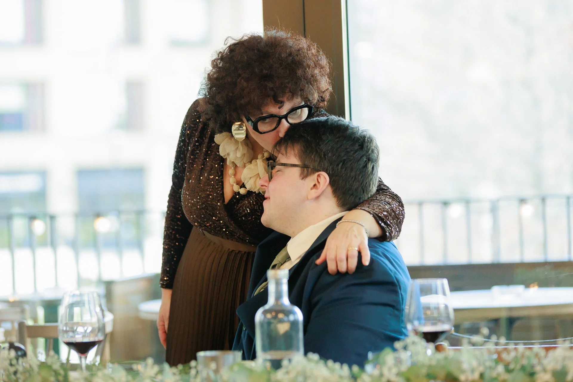 An older woman with curly hair, glasses, and large earrings gently kisses a young man with glasses on the forehead in a restaurant setting, both smiling with their eyes closed.