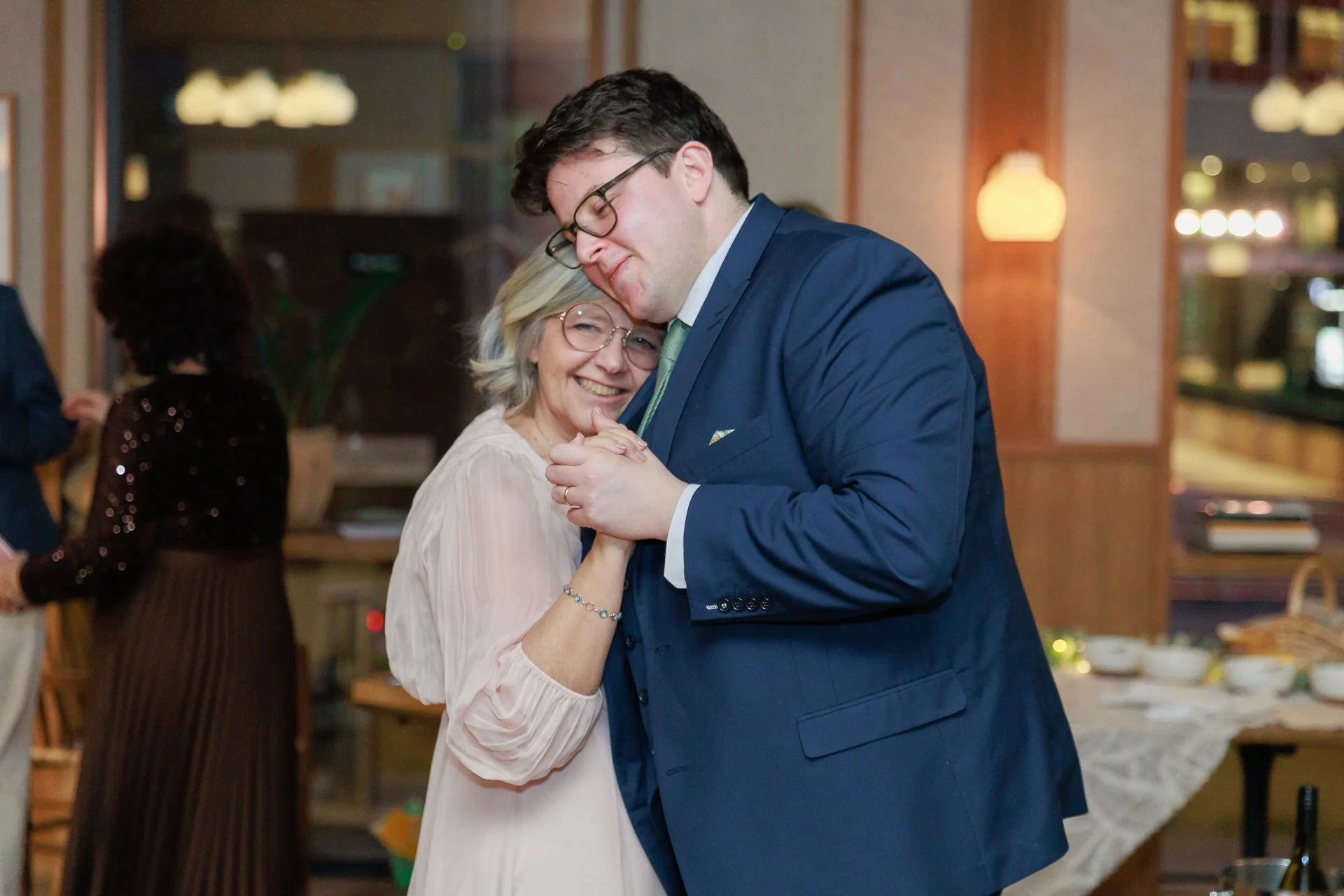 A young man in a blue suit and glasses dancing closely with an older woman with glasses and gray hair, both smiling warmly in a warmly lit room.